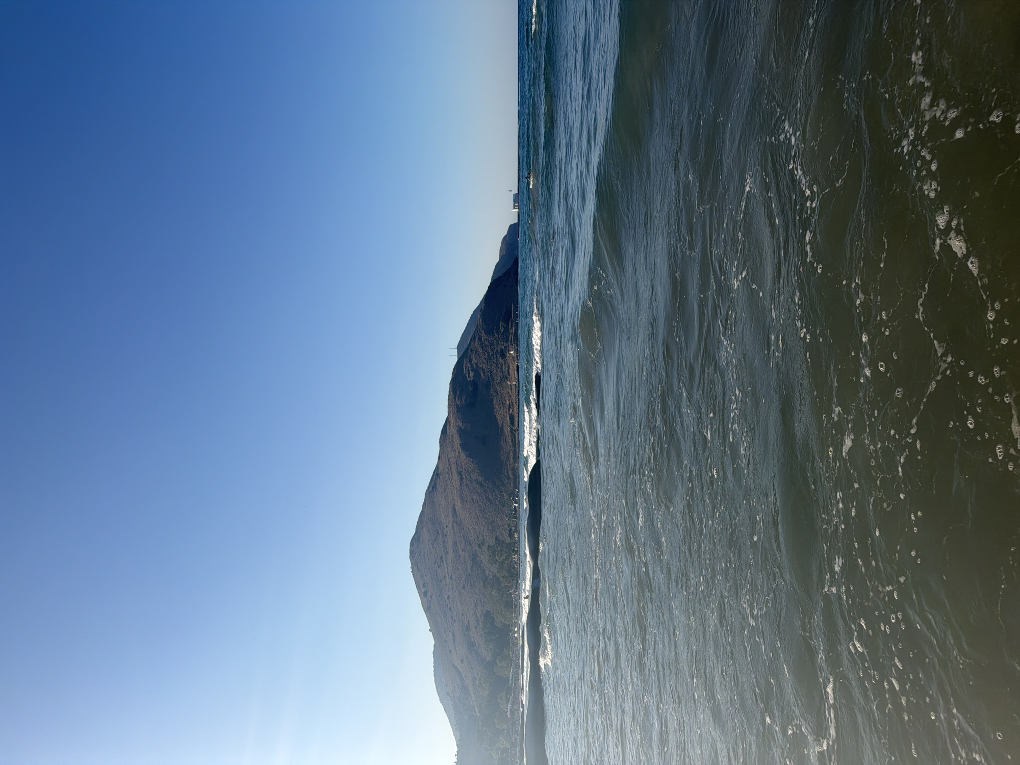 The image depicts a wide view of the sea with a mountainous coastline in the background, under a clear blue sky. The water in the foreground is choppy with small waves and white foam. In the middle ground, there are larger waves breaking, and a long, dark structure, possibly a breakwater or pier, runs horizontally across the frame. Beyond this structure, the water appears calmer. The coastline features a large, rounded mountain covered in vegetation, with some buildings visible at its base. Further to the right, along the coast, there is a white building, possibly a lighthouse, and a person on a paddleboard or surfboard. The scene is illuminated by sunlight, with a slight glare visible in the upper left corner of the sky, suggesting it is daytime. The location is identified as Georgioupoli, Greece, which is a coastal town known for its beaches and proximity to the mountains.