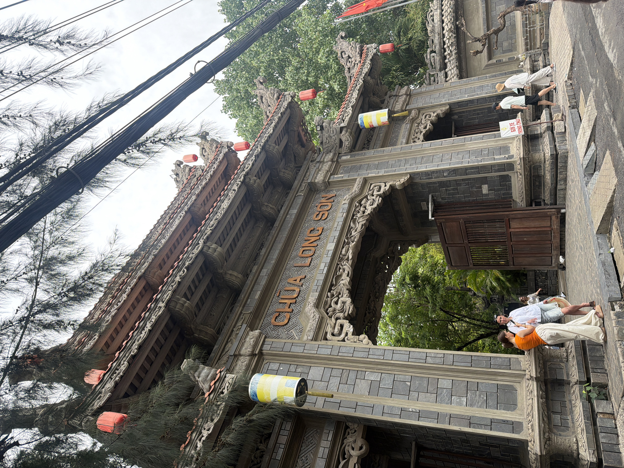 The image shows the entrance of Chua Long Son Temple in the city of Tay Nha Trang Ward, Vietnam. The temple's gate is a multi-tiered structure made of grey brick, intricately carved with traditional Vietnamese designs. Orange lanterns are suspended from the eaves of the temple's roof.
A few people are visible in the image. A couple in casual clothing stands near the entrance. Two other people are seen further in the background.
Electrical wires run across the upper left portion of the frame, contrasting with the traditional architecture of the temple. Trees are visible both in front and behind the temple structure.