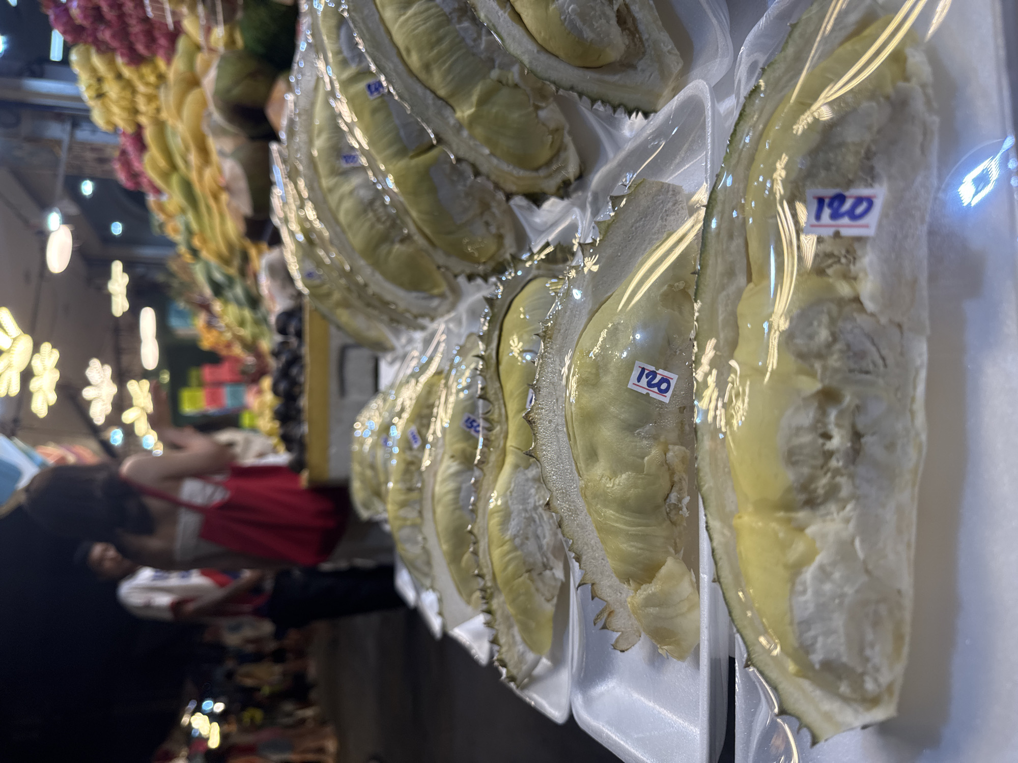 In Karon, Thailand, a nighttime fruit market stall displays several cut durian fruits for sale. The fruits are segmented and arranged on white styrofoam trays, each covered in clear plastic wrap. Small price tags, some reading "120," are affixed to the plastic. In the background, a blurry scene of people and stalls with illuminated lights suggests a bustling market environment.