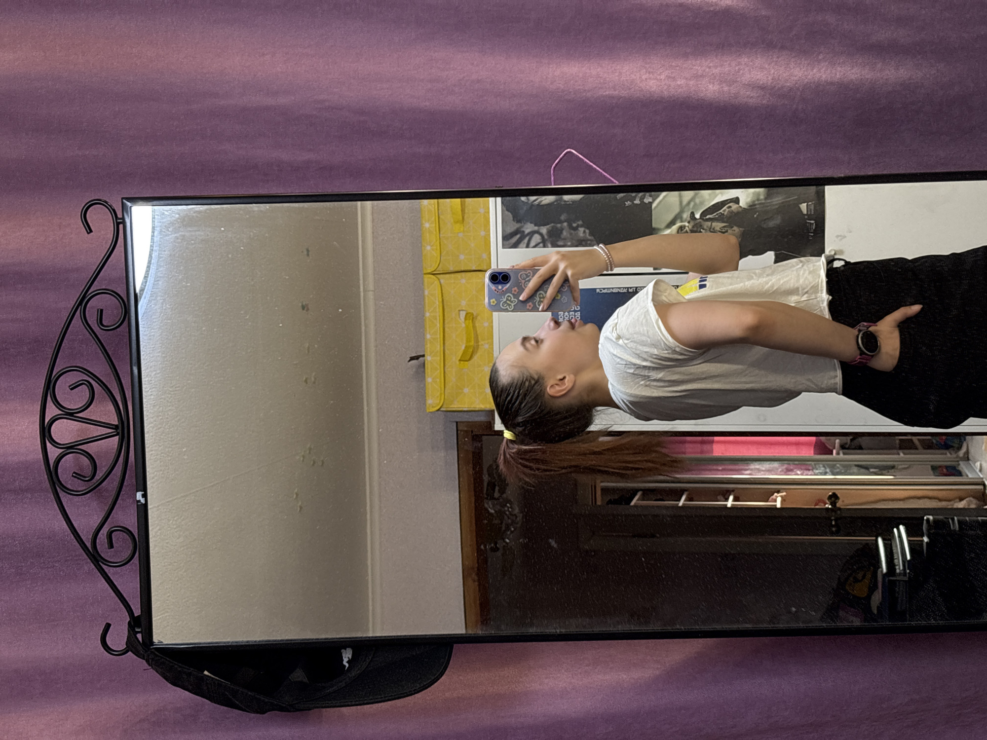 A mirror reflects a young woman holding a phone, with a purple surface partially visible behind it. The woman has brown hair tied back and is wearing a white shirt and black pants. The mirror has a black decorative frame with ornate scrollwork on one side. The reflection also reveals items on shelves and a poster on the wall. The scene appears to be indoors. The city is not discernible from the image.