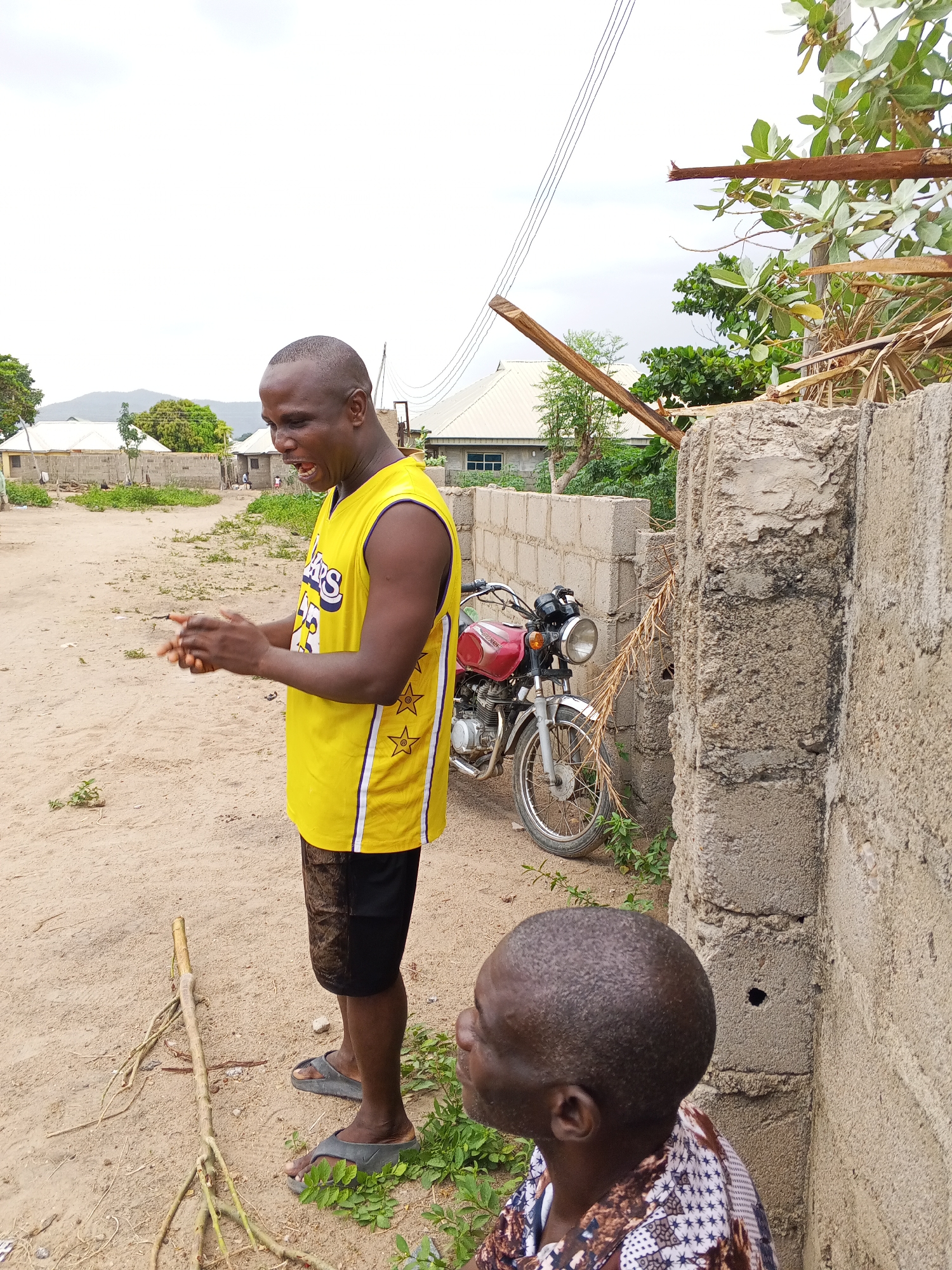 This outdoor image, taken in Zango-ndaji, Nigeria, features two men amidst a semi-rural setting. The central subject is a man with dark skin and short hair, standing and facing left. He is wearing a vibrant yellow sleeveless Los Angeles Lakers jersey with black shorts and grey slide sandals. His mouth is open, suggesting he is speaking, shouting, or laughing, and his hands are held together in front of him, possibly gesticulating or clapping.
Another man, also with dark skin and short hair, is visible in the foreground, crouched or seated low, with only the back of his head and shoulder visible. He wears a patterned white and brown shirt and appears to be looking up and to the left, towards the standing man or something in that direction.
The setting consists of a sandy or dirt ground with sparse green vegetation. A red motorcycle with chrome accents is parked behind the standing man, leaning against a concrete block wall. More similar concrete walls and fences are visible, delineating parts of the area. In the background, several simple houses with light-colored roofs are scattered, and a distant mountain range is faintly visible under an overcast, bright sky. Overhead, power lines stretch across the frame, and broken tree branches are evident, some resting on the walls. The time of day appears to be daytime, likely late morning or afternoon, given the diffused light. The visible text on the standing man's jersey clearly reads "Lakers" on the front and features star patterns on the side panels.