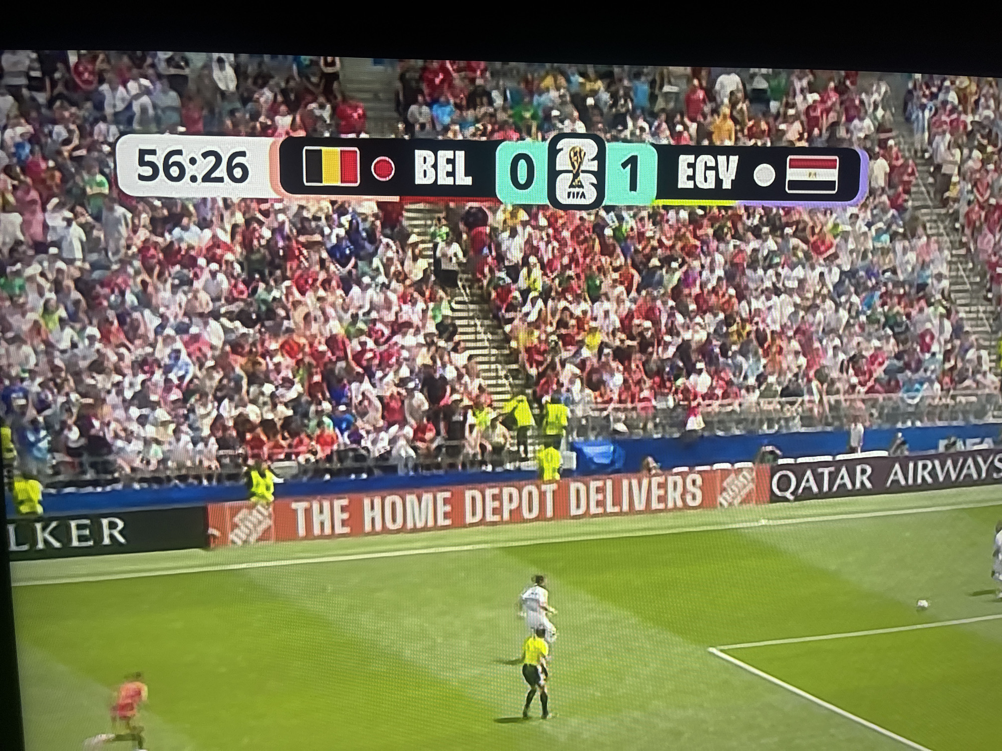 The image is a screenshot of a soccer match broadcast, showing a scoreboard and a portion of the stadium seating filled with spectators. The scoreboard displays the game time as 56:26, with the score showing Belgium (BEL) with 0 goals and Egypt (EGY) with 1 goal. The Belgian flag is shown next to "BEL," and the Egyptian flag is next to "EGY." The FIFA World Cup logo is also visible on the scoreboard. The stadium seats are packed with people wearing various colored clothing, predominantly red, white, and blue. In the foreground, there is a green soccer field with several players. Two individuals in yellow vests, likely stadium staff, are standing near the edge of the field. Additional advertising banners are visible along the side of the field, including "LKER," "THE HOME DEPOT DELIVERS," and "QATAR AIRWAYS." The image does not provide any information about the location being the city of Ruse, Bulgaria.