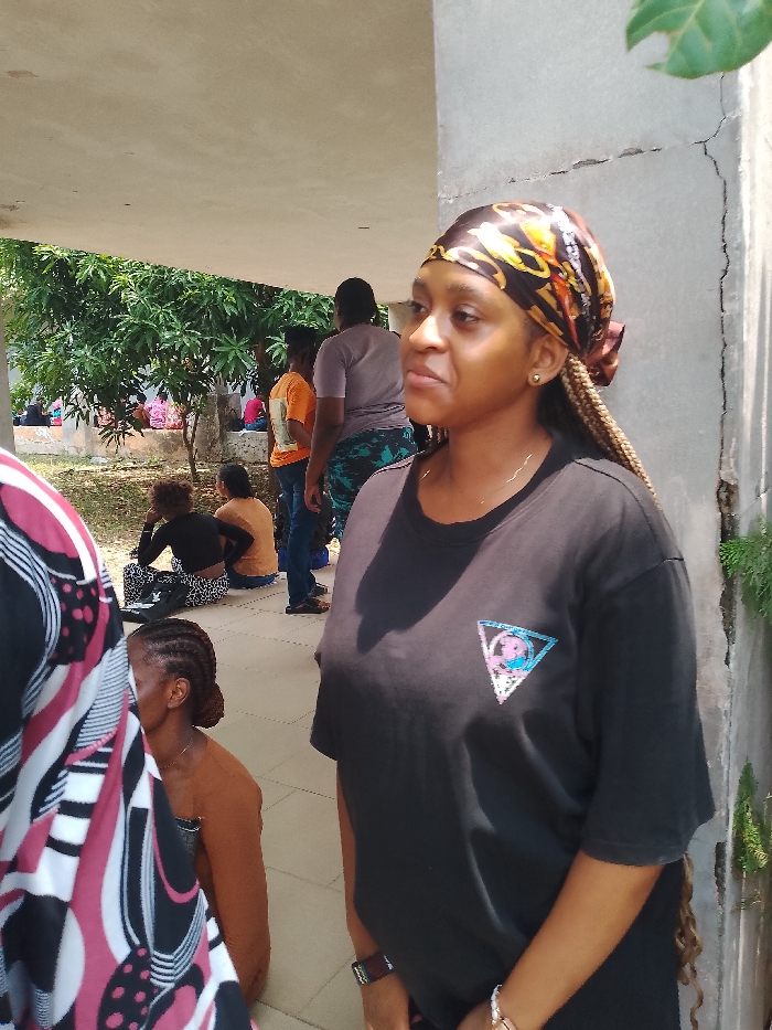 The image depicts a group of people in an outdoor setting, possibly within the city of Urualla, Nigeria. A woman with braided hair, wearing a black t-shirt and a patterned headscarf, stands prominently in the foreground, looking towards the right. In the background, other individuals are seated or standing, some appearing to be waiting. The location seems to be a covered area, possibly a building or a structure with a roof, adjacent to some trees and vegetation. A concrete pillar with cracks is visible on the right. The scene suggests a casual gathering or waiting area.