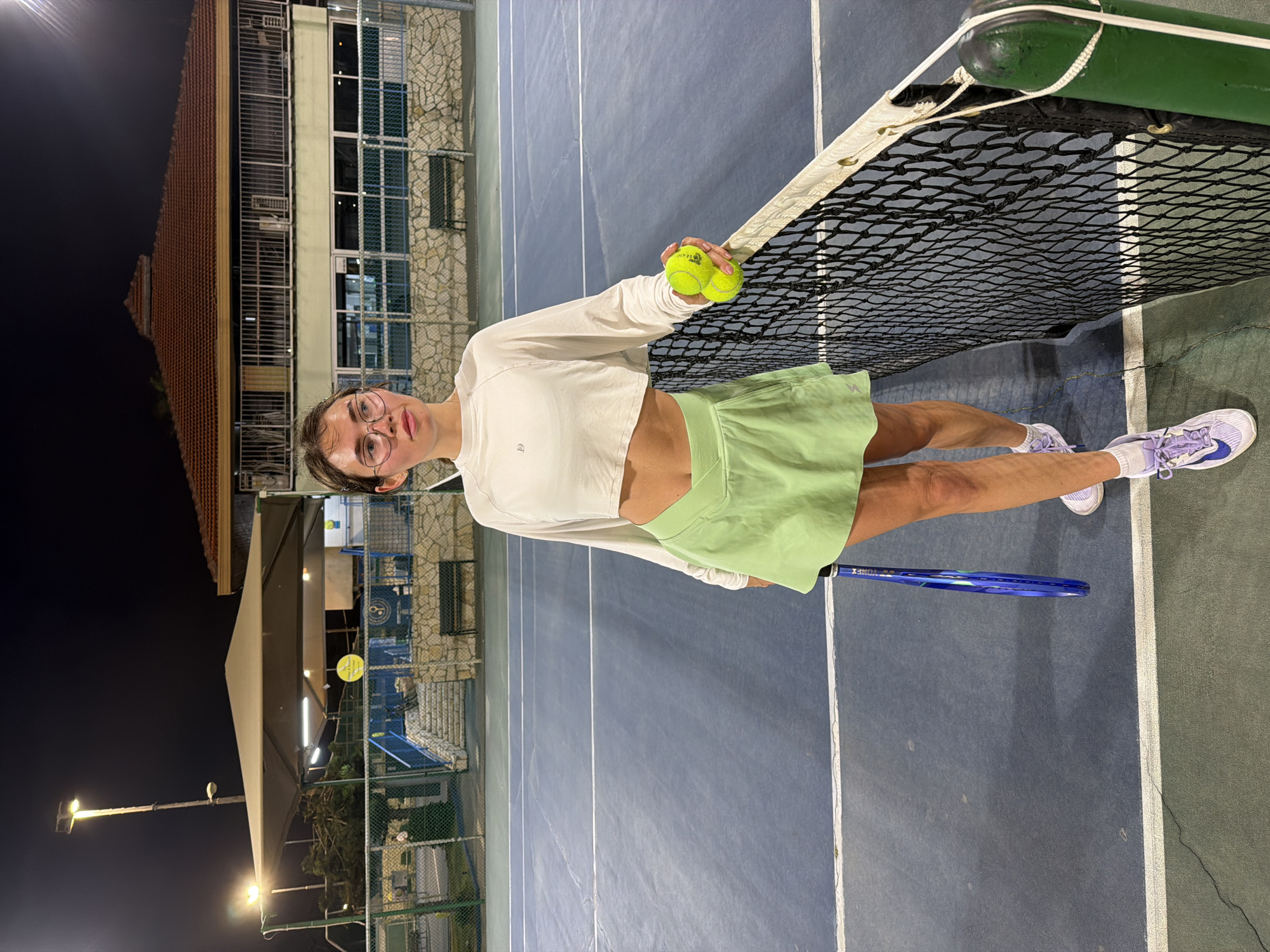 A full shot captures a young adult standing on a blue and green tennis court outdoors. The person is light-skinned with brown hair pulled back, wearing round glasses, a long-sleeved white cropped top, and a light green tennis skirt. The person is holding two yellow tennis balls in their right hand.
To the right of the person, the tennis net stretches across the court. A blue and green tennis racket is at the person's left side. The person wears white sneakers with purple accents.
The background features the blue tennis court with white lines, with a multi-story building with orange roofs on the left, and floodlights illuminate the tennis court. A building with a stone facade is visible behind the court. The image was taken at night.
The location of the scene is likely a tennis court in Tel-Aviv, Israel.