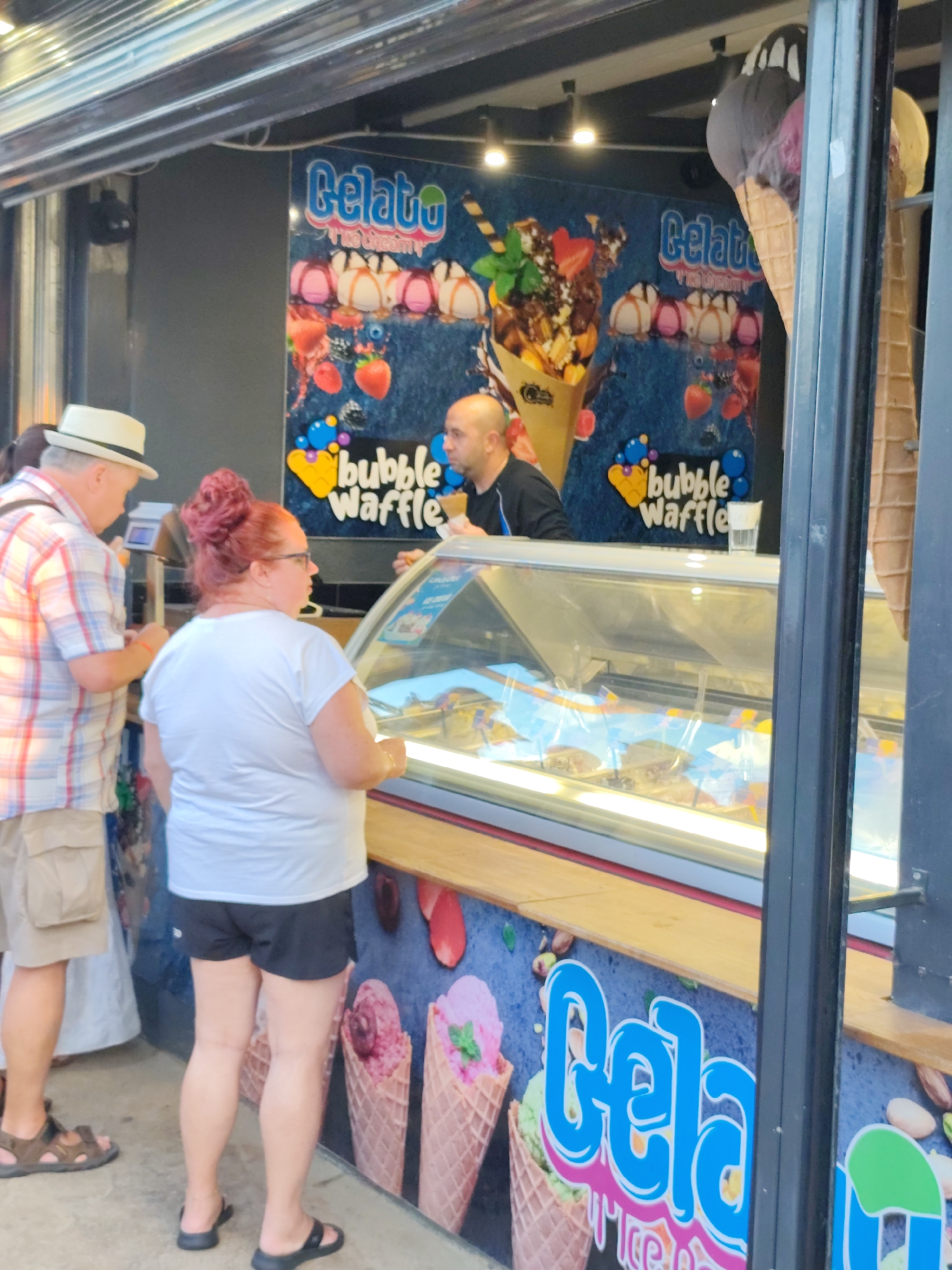 An outdoor ice cream stand in Nesebar, Bulgaria, features a bright blue and pink "Gelato" sign and a mural depicting various ice cream cones and toppings. Three people are visible: a man in a checkered shirt and a hat, a woman with red hair pulled up in a bun wearing glasses and a white t-shirt, and a man in a black shirt. The woman appears to be ordering from the ice cream display case. The ice cream stand has a refrigerated display case filled with various flavors of ice cream. The background shows a textured wall with the ice cream advertisements. The lighting suggests it is daytime, possibly late afternoon or evening, with some artificial lighting from the stand.