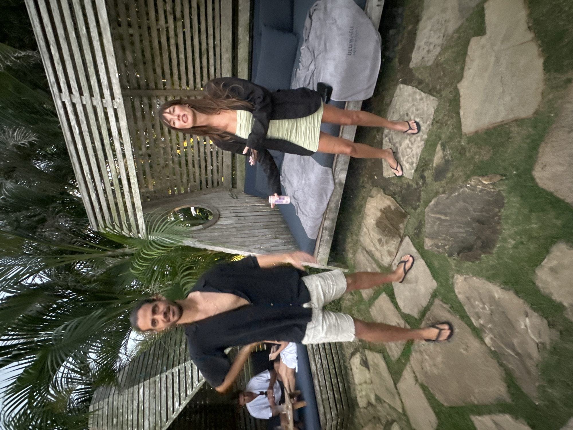 Two adults, a man and a woman, stand in a paved outdoor area with grass and large stones. The man, on the left, wears an unbuttoned black short-sleeved shirt and light-colored shorts. He has dark hair pulled back and is looking at the camera. The woman, on the right, wears a green strapless dress and a dark blazer. She is holding a pink can and looking to her left with her mouth open. Behind them is a wooden structure with slatted walls and a cushioned seating area covered by gray blankets. In the background, another person is seated on a lounge. The environment appears to be a tropical setting with palm trees visible overhead. This scene is consistent with the Uluwatu area of Kuta Selatan, Indonesia, known for its resorts and beach clubs.