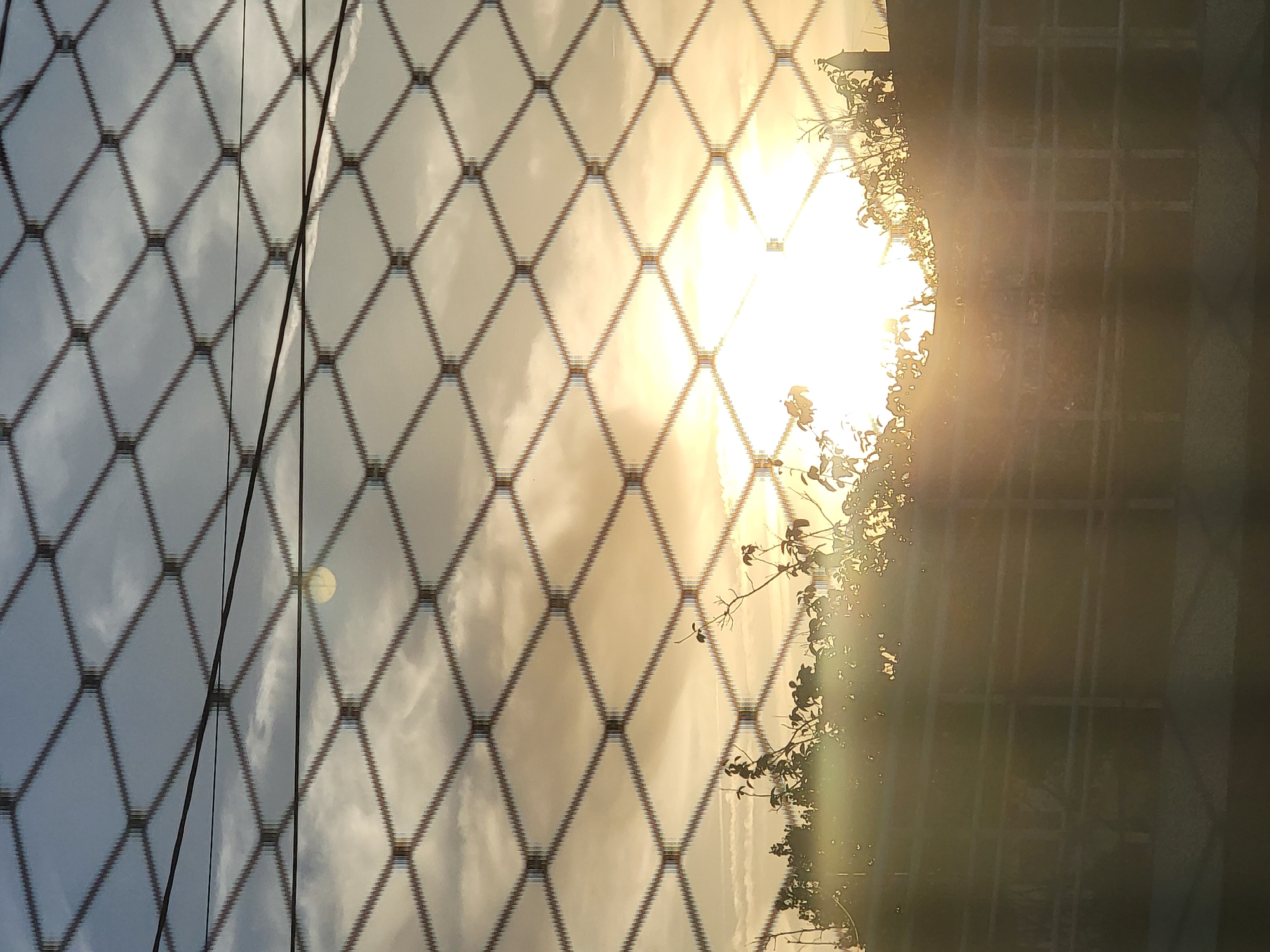 The image depicts a sunrise viewed through a chain-link fence. The sun is a bright, washed-out orb positioned towards the right side of the frame, casting a strong glare. Silhouetted against the sky are the branches of trees and foliage, as well as what appears to be a low wall or structure. The sky itself is a mix of pale blue and warm, hazy tones, with wispy clouds. A power line runs horizontally across the upper portion of the image, and the texture of the chain-link fence is prominently visible, creating a diamond-shaped pattern that overlays the entire scene. The overall impression is one of a typical urban or suburban sunrise in Faro, Portugal, seen from a vantage point behind a barrier.