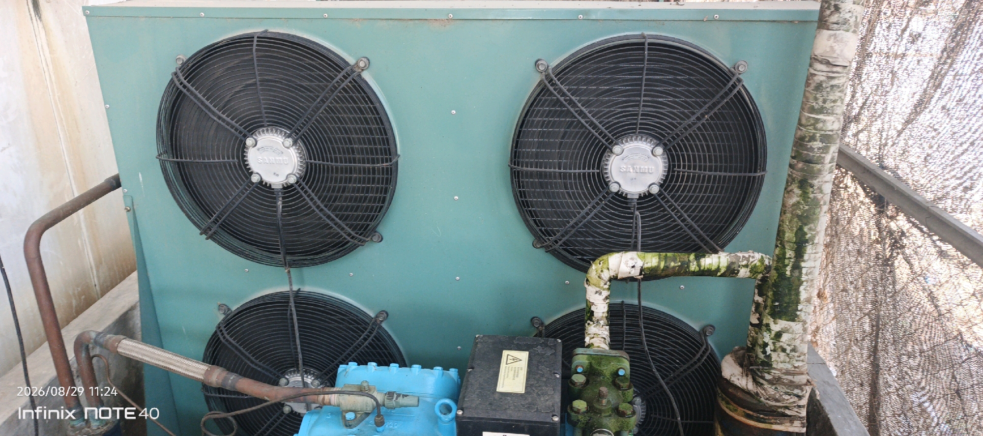 An outdoor air conditioning unit with four large fans is visible in the city of Patampanua, Indonesia. The unit is teal green and has the brand "Sanmu" visible on the center of each fan. To the right of the unit, a tree trunk with peeling bark and moss is present, with a metal support beam running vertically behind it. In the background, a dark mesh screen is stretched taut. Copper pipes are connected to the unit, one of which appears to be a flexible metal hose. A blue compressor is also visible at the bottom of the unit, with a black electrical box attached to it that has a yellow warning sticker. The timestamp "2026/08/29 11:24" and "Infinix NOTE 40" are overlaid on the bottom left corner of the image.