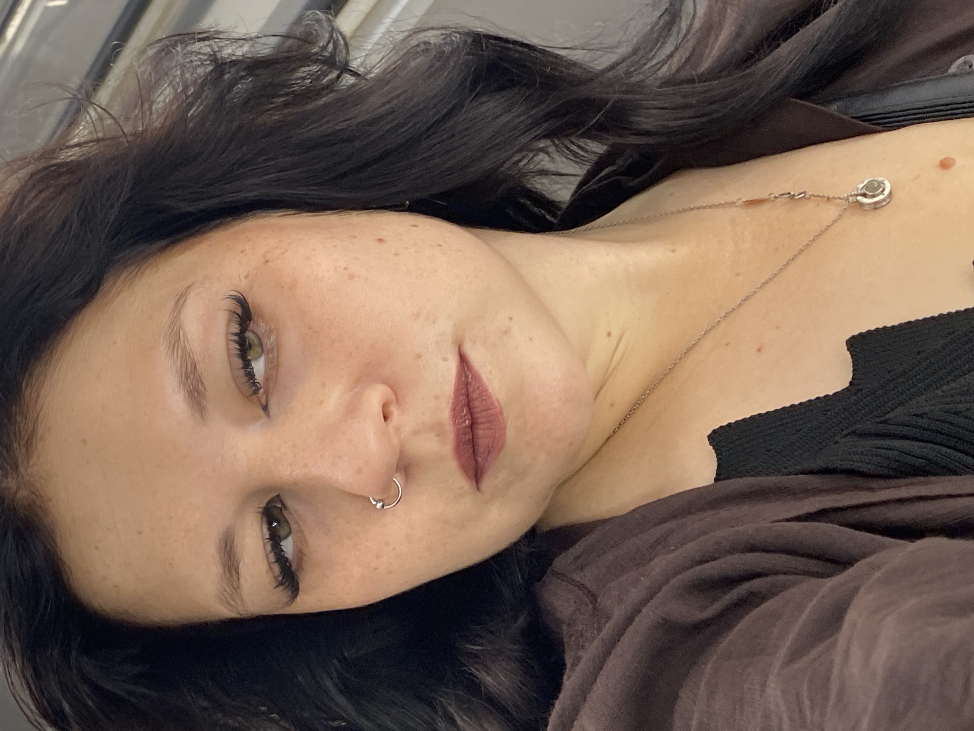 A close-up, head-and-shoulders shot shows a woman with dark, wavy hair and light brown eyes. She is wearing a brown open-fronted top over a black ribbed tank top and a silver necklace with a circular pendant. She has a silver hoop nose ring and appears to be wearing false eyelashes. Her lips are painted with a matte, brownish-red lipstick. The background is blurred and appears to be indoors with artificial lighting. There is no discernible information in the image to suggest a specific location in Saint Petersburg, Russia.
