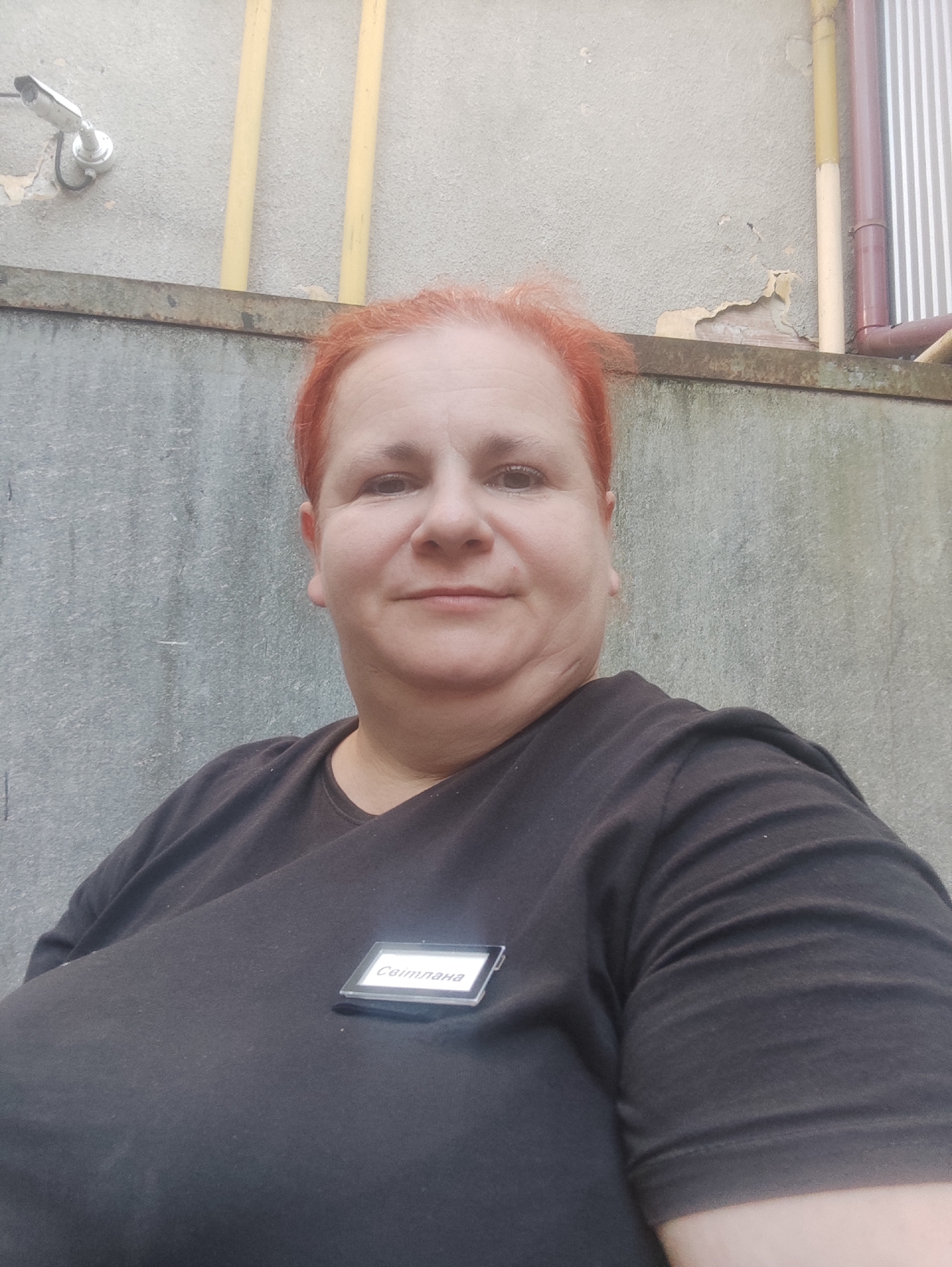 A medium shot captures a woman with short, red hair and fair skin, facing the camera and smiling slightly. She is wearing a black t-shirt with a name tag that reads "Світлана" affixed to the left side of her chest. The background consists of a textured grey concrete wall and a section of a lighter grey concrete structure above it. To the left of the frame, a white security camera is mounted on the upper wall. Two yellow pipes and a segment of a pipe with vertical white stripes are visible on the right side of the upper wall. The overall environment appears to be an outdoor, possibly urban, setting.