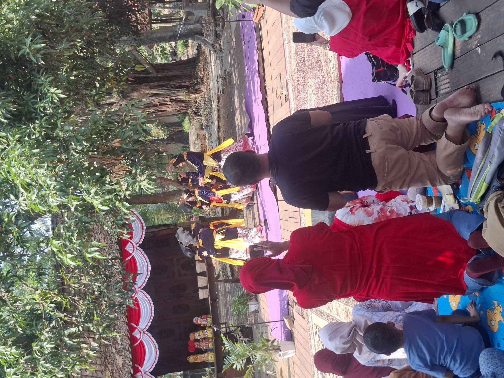The image captures an outdoor cultural performance, likely a traditional dance, taking place on a wooden stage covered with a purple mat. Several individuals, dressed in vibrant traditional attire including yellow sashes and patterned skirts, are actively performing on the stage. In the foreground, a seated audience observes the performance. One person is kneeling, facing the stage, while others are also seated, some with heads covered. The backdrop features a structure with a red and white decorative curtain, suggesting a temporary stage setup. Lush green trees with dense foliage dominate the upper portion of the image, indicating a natural or semi-rural environment. The overall scene suggests a community event or cultural exhibition possibly held in a public space within the city of South Tangerang, Indonesia, given the characteristic tropical foliage and architecture often found in such locations.
