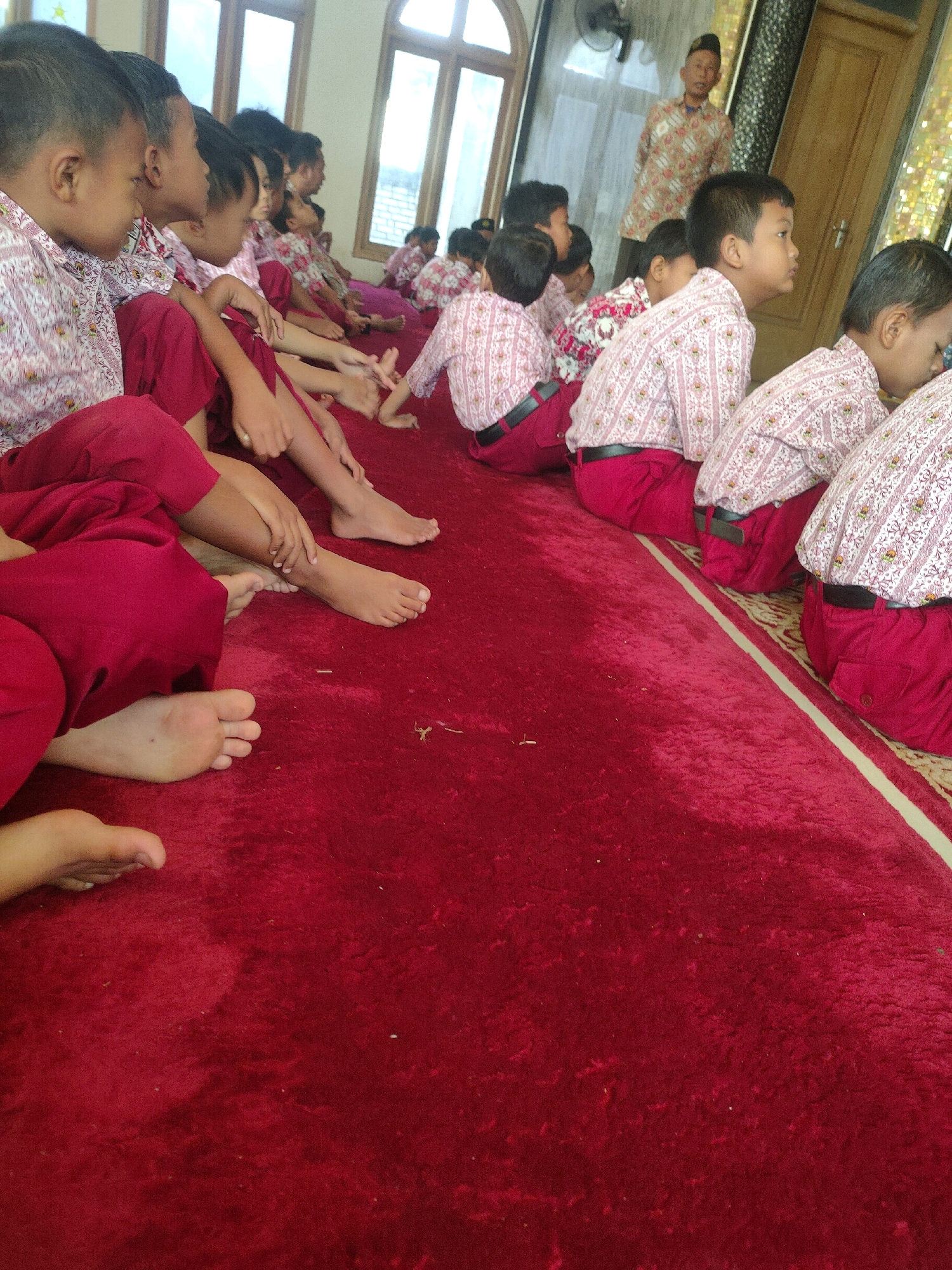 In Payaman, Indonesia, a group of young boys, likely students, are seated on a plush red carpet. They are all wearing similar uniforms: white shirts with a subtle pink pattern and red pants. Some of the boys are looking towards the right of the frame, where a man in traditional Indonesian attire stands. The environment appears to be indoors, with large windows providing natural light. The overall scene suggests a formal gathering or a lesson.