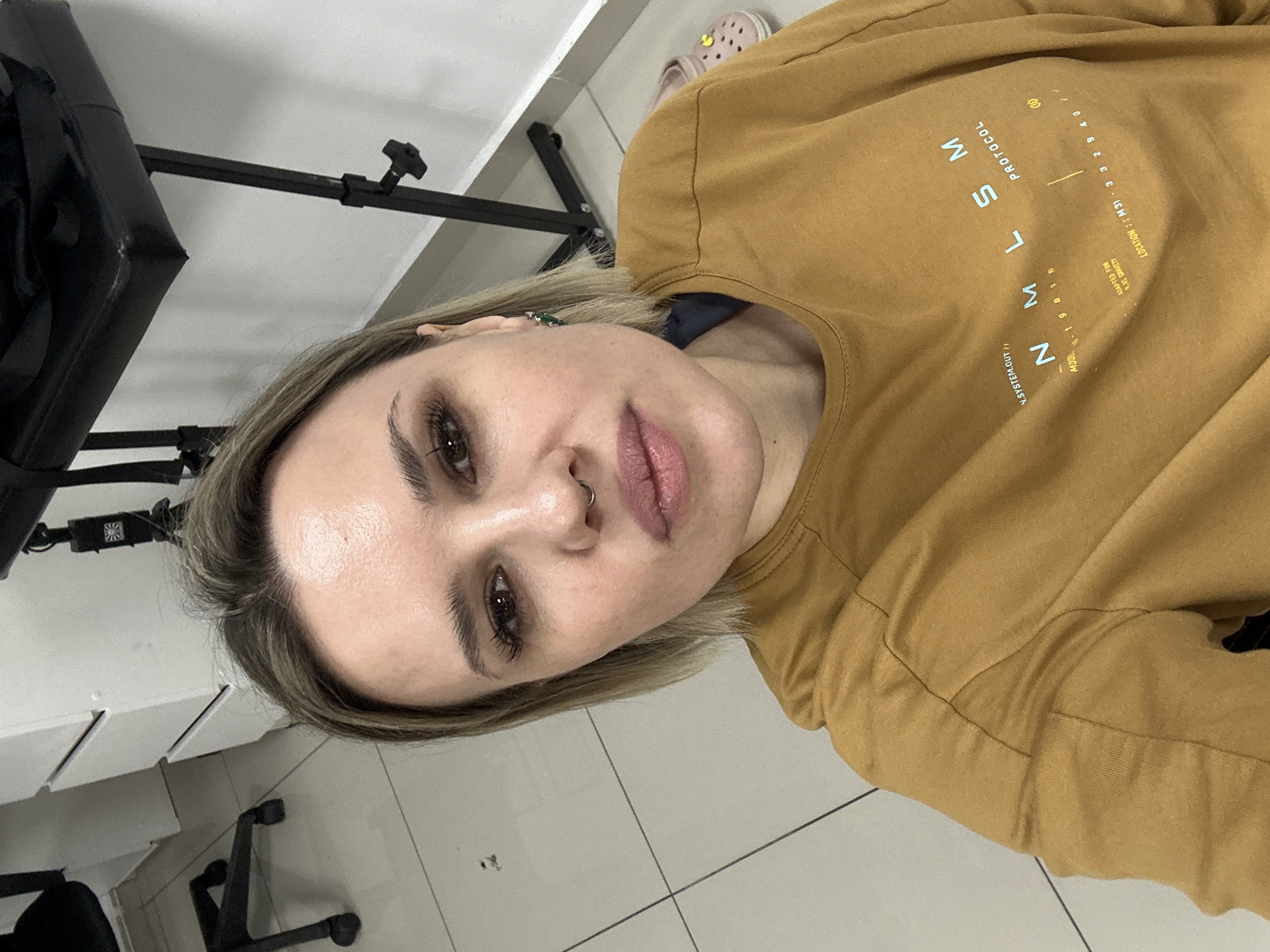 A selfie of a woman with short, light brown hair and a nose ring, wearing a mustard-colored sweatshirt with printed text. The text includes "N.SYSTEM.OUT", "PROTOCOL" and numbers/coordinates. The background includes a white tiled floor, the leg of a black chair, part of a white desk or cabinet, and a black object on a stand (likely a light or equipment). The location and city are not discernible from the image.