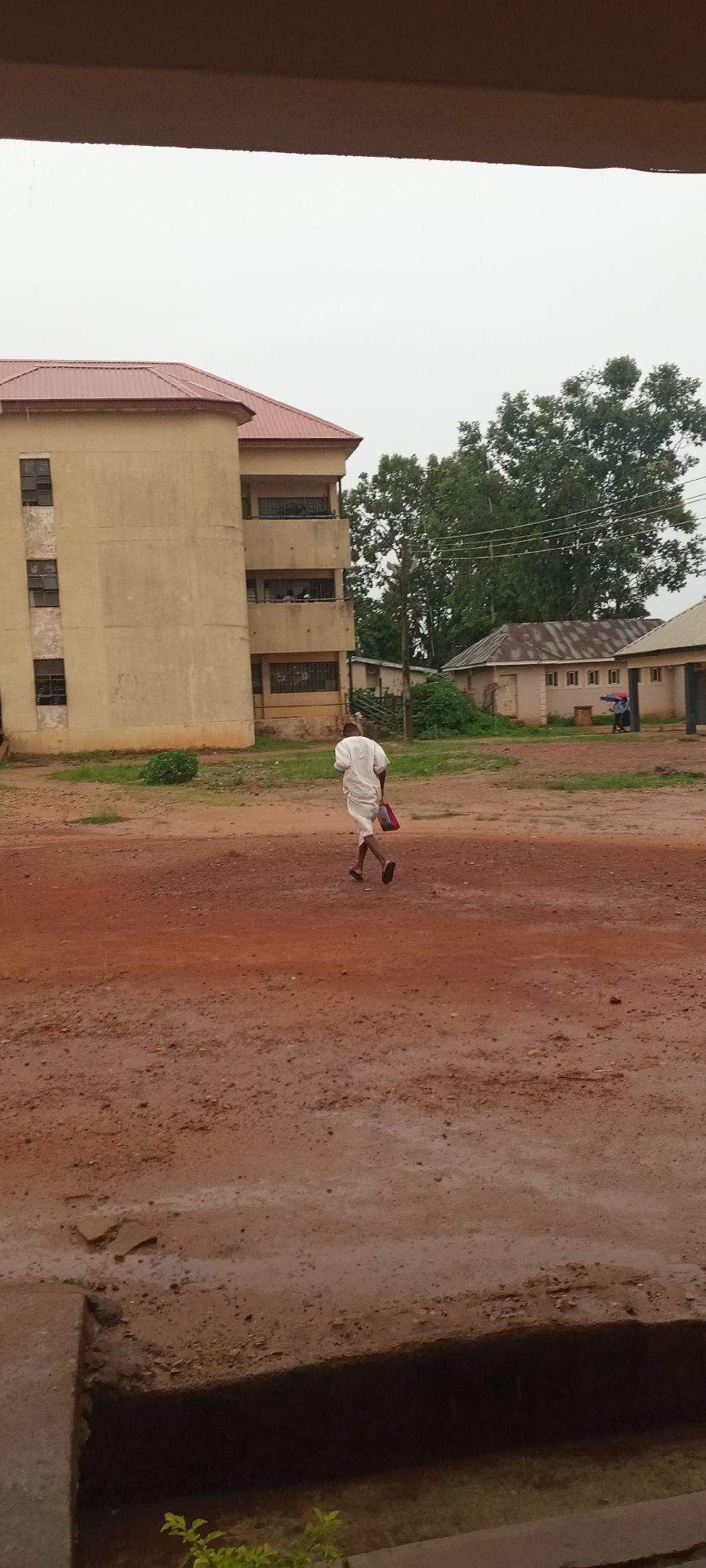 A medium shot, taken through an open window, depicts a man walking across a muddy, reddish-brown terrain towards a building complex. The man is clad in a white garment and carries a red bag. Behind him stands a three-story building with a light-tan exterior and a red roof. Adjacent to it is a one-story building with a corrugated roof. A mature tree with dense green foliage is visible in the background, partially obscuring the second building. The sky appears overcast, and the scene is generally dimly lit.