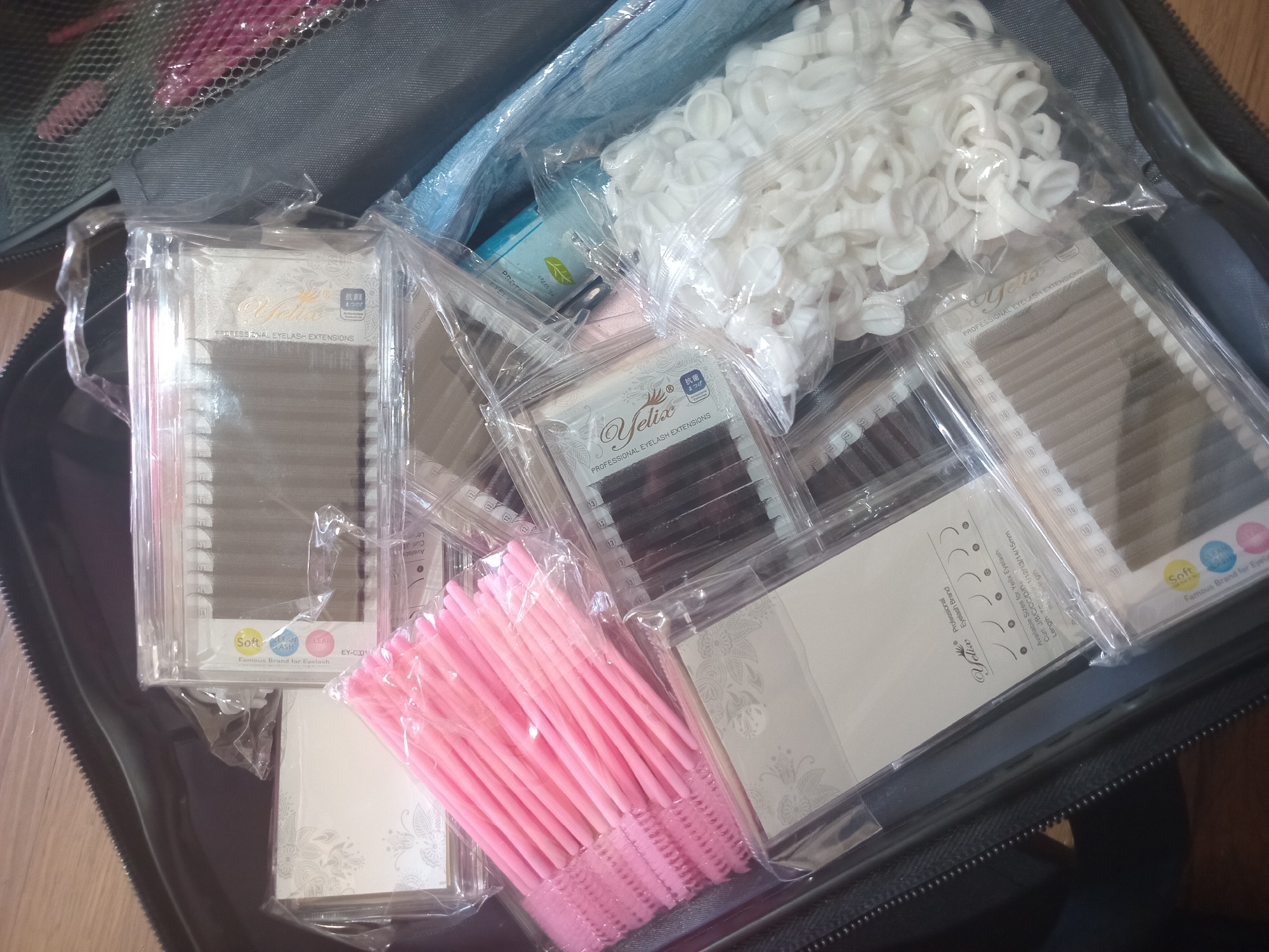 An overhead shot captures a collection of eyelash extension supplies neatly packed inside a dark, zippered soft-sided case. The main subjects are numerous clear plastic trays containing professional eyelash extensions, predominantly featuring the "Yelix" brand and also "Famous Brand for Eyelash." Many trays show individual lash lengths, such as '10'.
Among the various items, a large bag filled with white, flower-shaped adhesive rings/cups is visible in the upper right. Below it, a plastic bag holds numerous pink disposable mascara wands or brushes. A small blue package, partially obscured, shows text including "PROTEIN" and "GREEN TEA."
Visible text on the eyelash extension packaging includes "Yelix," "PROFESSIONAL EYELASH EXTENSIONS," "Soft," "LEAN," and "Famous Brand for Eyelash." Specific details like "Curl: J/B/C/CC/D/D+" and "Length: 1/12/13/14/15mm" are discernible on some labels. Japanese characters like "抗菌" (antibacterial) are also present. The scene is indoors, likely on a wooden surface, and depicts tools and supplies for beauty services, suggesting preparation for, or organized storage after, an eyelash extension application session. No people are visible, and no specific time of day or weather can be determined. The image provides no visual cues to confirm the external location context of North Kuta, Indonesia.