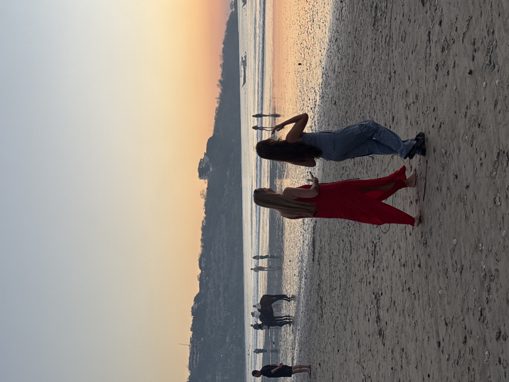 Two women walk on a wet sandy beach during sunset. The woman on the left wears a long, flowing red dress with a slit up the side. The woman on the right wears a blue denim jumpsuit. They are walking away from the camera towards the ocean. In the middle ground, several people are standing in the shallow water. A horse is also present in the water, with a person on its back and another person standing next to it. Further out in the ocean, there are two boats. The background features a coastline with vegetation and buildings, silhouetted against a gradient sky of orange and blue. The overall scene is tranquil and evokes a sense of a peaceful evening at the beach.