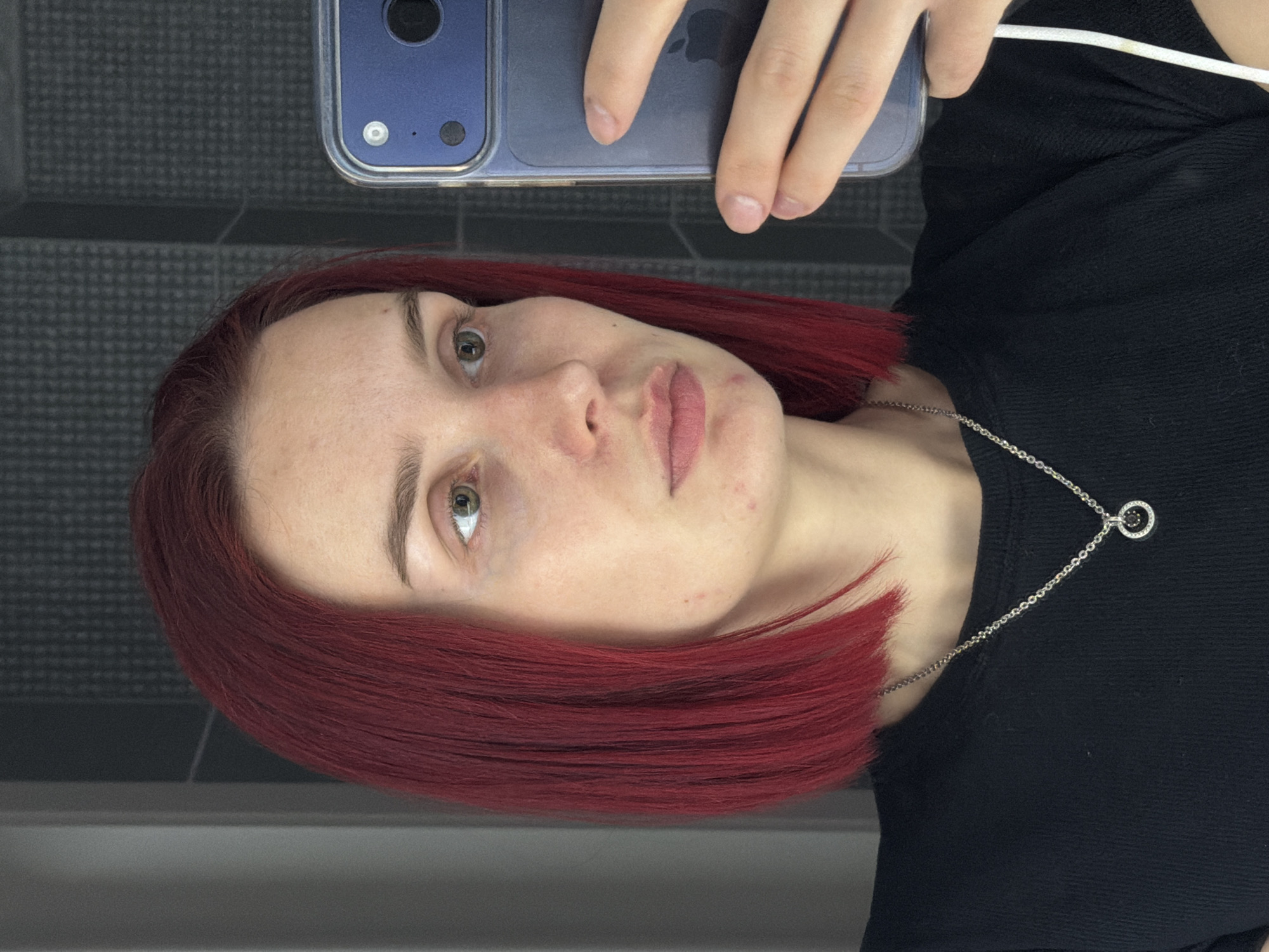 A young woman is captured in a selfie taken in Chaikovsky, Russia. Her head is tilted, and she looks directly into the camera with a neutral expression. She has striking, vibrant red hair that frames her face, light skin, and green eyes. Minor blemishes are visible on her forehead and cheeks. She wears a plain black top and a silver chain necklace with a small, circular pendant featuring a dark center. She is holding a blue-hued smartphone, encased in clear plastic, with her hand visible at the top of the frame. A faint Apple logo can be seen on the back of the phone, and a white cable extends from its upper right side. The background features a dark gray tiled wall with a grid pattern, below which runs a dark horizontal band, suggesting an indoor environment. No specific time of day or strong emotions are evident.