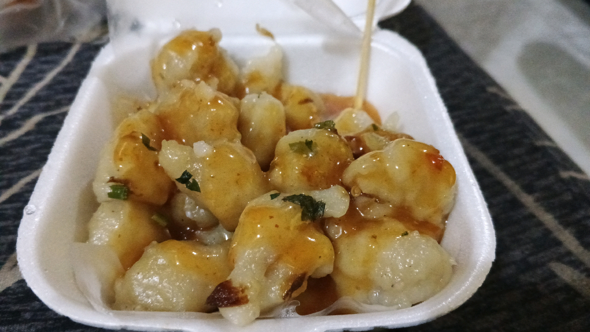 A close-up, eye-level shot shows a white styrofoam container filled with a savory dish. The food consists of small, light-colored dumplings or meatballs, coated in a glistening, orange-brown sauce. Some of the dumplings have visible green specks, likely herbs or spices. A wooden skewer is inserted into some of the food items, and a thin stream of the sauce is seen being poured over the top from an unseen source above. The styrofoam container is partially filled, allowing the dark, patterned background of a table or surface to be visible around the edges. The lighting is somewhat dim, highlighting the glossy texture of the sauce. The image is focused on the food, with the background slightly blurred. This dish is likely being served as takeout or a casual meal.