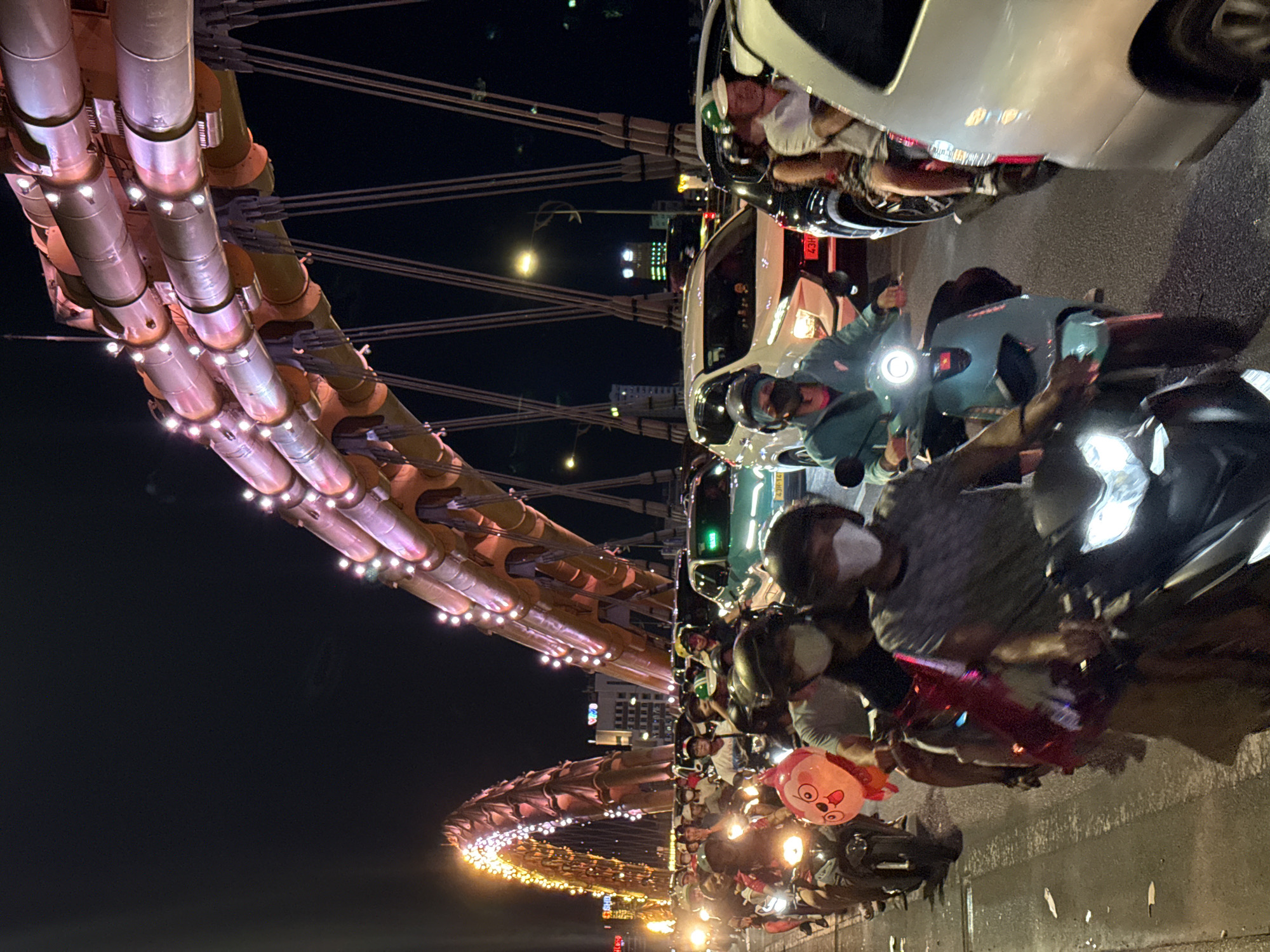The image captures a densely trafficked scene at night on the Dragon Bridge in Đà Nẵng, Vietnam. The prominent arch of the Dragon Bridge is illuminated with numerous bright lights, showcasing its segmented, pipe-like structure and suspension cables. In the background, a second, similarly illuminated arch of the bridge is visible.
The roadway is packed with vehicles, primarily motorbikes and several cars, suggesting heavy traffic flow. Numerous individuals are seen riding motorbikes, many wearing helmets and some sporting white face masks. A distinctive red, round balloon with a cartoonish face is attached to the rear of a motorbike in the left foreground. Car headlights and motorbike headlamps are illuminated, casting light on the immediate surroundings.
Among the cars, a white SUV is visible in the middle ground, and a teal-colored car, possibly a taxi given a discernible logo on its rear window, is also present. Overhead streetlights emit a yellowish glow, and distant urban buildings with illuminated windows are faintly visible against the dark night sky. The scene depicts an active urban environment during evening hours.