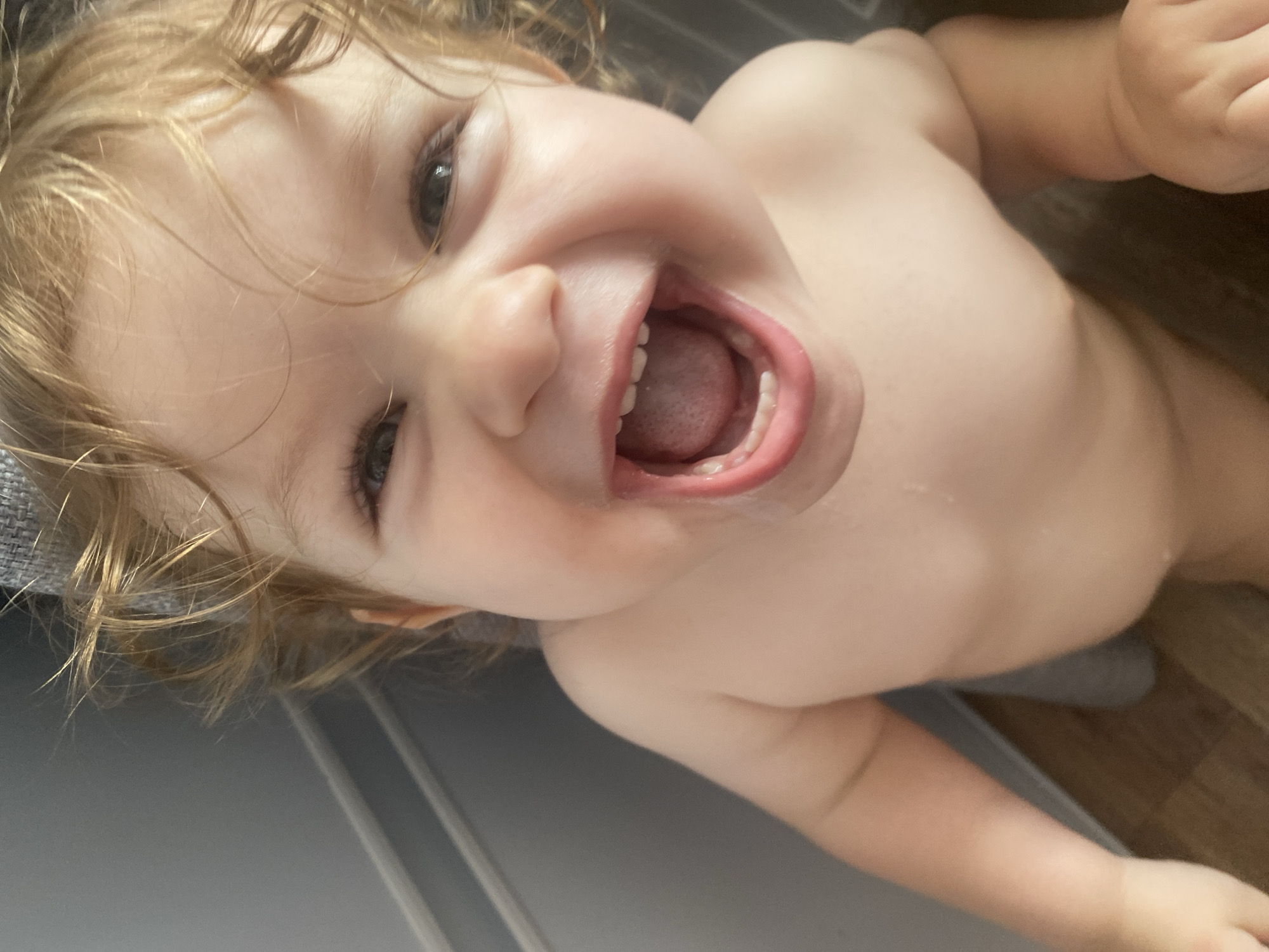 An extreme close-up, low-angle shot captures a young Caucasian child with curly blonde hair, bare-chested and laughing with their mouth wide open. The child's eyes are visible and appear to be looking upwards. The background is out of focus, showing a gray wall with a horizontal molding, and a dark floor. The lighting is bright, illuminating the child's face.