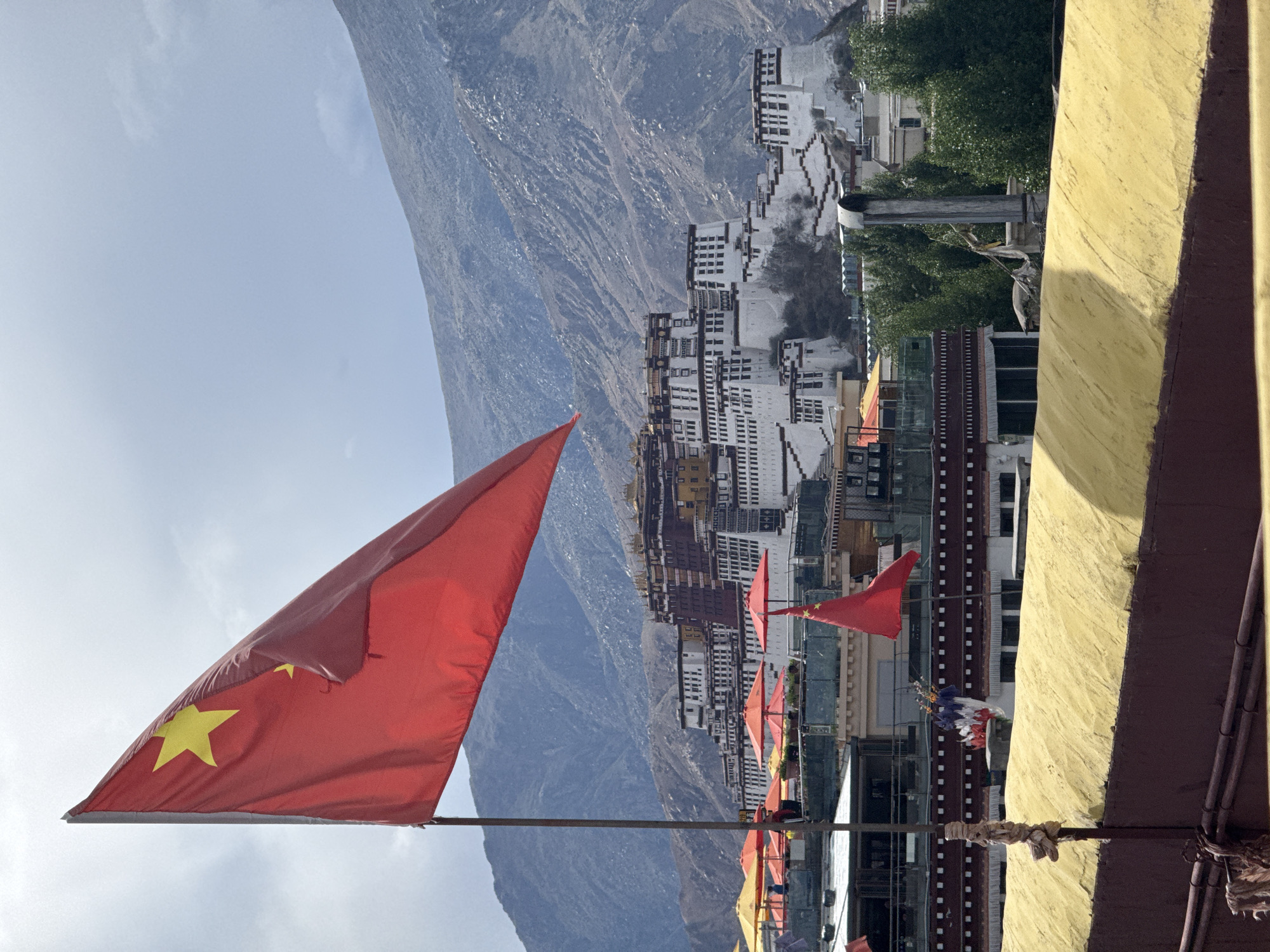 This elevated shot captures a panoramic view of the majestic Potala Palace in Lhasa, Tibet, China, under a bright, clear sky. In the immediate foreground, a large, vibrant red Chinese flag proudly billows, displaying its prominent five yellow stars – one large star accompanied by four smaller ones. Its flagpole anchors it to the bottom-left of the frame, setting it against the expanse of the sky.
Further into the mid-ground, a series of smaller red flags, also featuring yellow stars, are strung along what appears to be a railing or wire, contributing to a celebratory or official ambiance. These flags guide the eye towards the iconic Potala Palace, which stands as the central subject.
The Potala Palace complex is a striking multi-story structure, characterized by its distinct white and reddish-brown sections, meticulously built into the natural contours of a hill. The intricate details of its numerous windows and tiered architecture are discernible, hinting at its historical grandeur. Scattered among the palace grounds and surrounding buildings are patches of green foliage, indicating trees. On the right side of the frame, a weathered yellow wall forms part of the vantage point from which the photograph was taken.
In the distant background, formidable, rugged mountains extend across the horizon, complementing the high-altitude setting. The bright sunlight suggests the image was captured during the day, likely late morning or early afternoon, with only a few faint clouds in the otherwise clear sky. The image powerfully combines natural landscape, significant architecture, and national symbolism. No people or visible text are present in the scene.