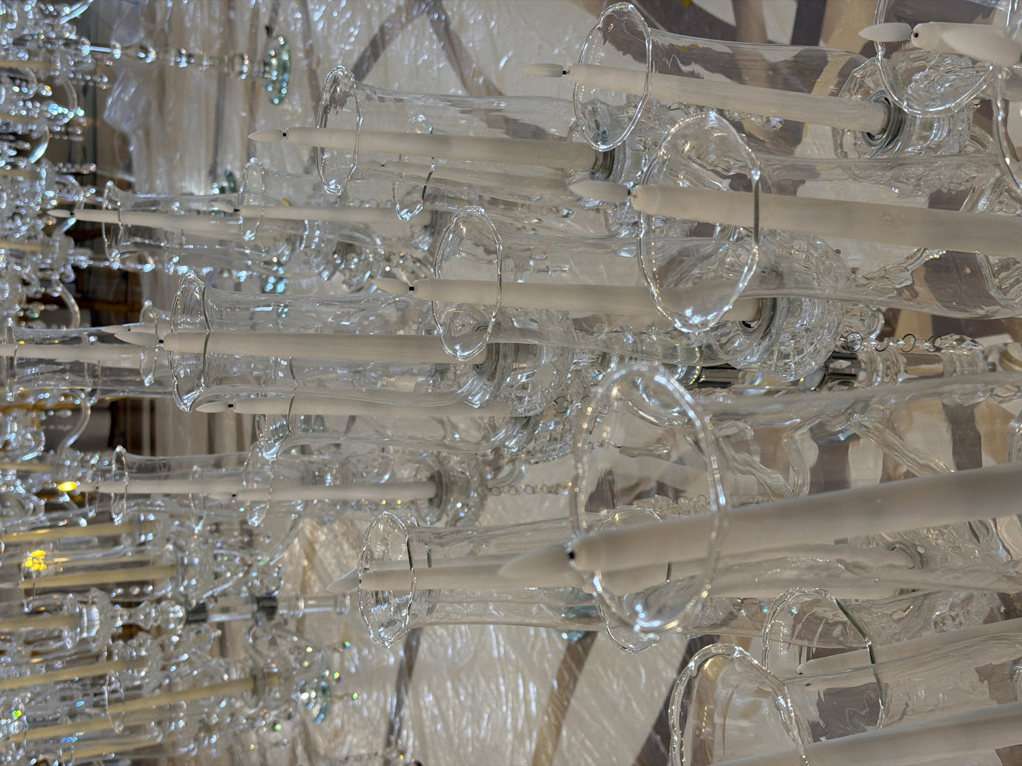 The image shows a chandelier in the city of Moscow, Russia. The chandelier is made of clear glass, and it is in a horizontal position. It has many long white candles. The background appears to be a fabric covering.