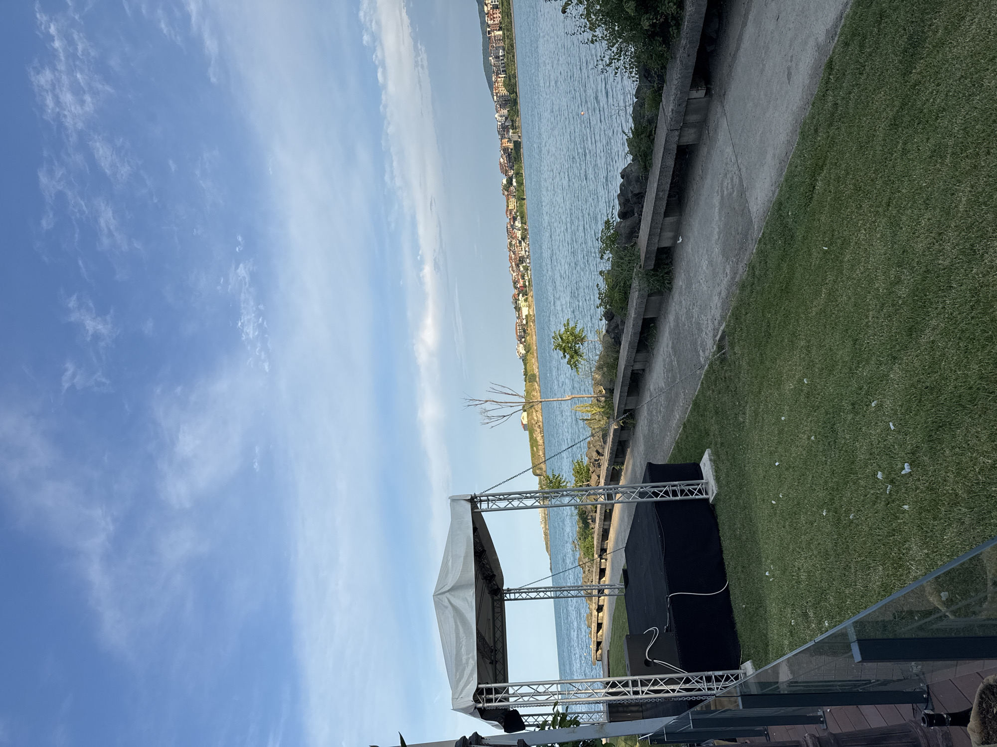 The image depicts an outdoor coastal scene in Tsarevo, Bulgaria, under a blue sky with scattered white clouds. In the foreground, a temporary event stage with a gray canvas canopy supported by four metal truss pillars and a black platform is visible on a green lawn. White cables and black audio equipment are present on the stage. The lawn is sparsely dotted with small, white, feathery objects. A clear glass railing separates the lawn from a partially visible wooden deck on the left.
To the right of the stage and lawn, a concrete walkway runs parallel to the water's edge. Along this walkway, multiple long, grey concrete benches are positioned facing the water. Between the walkway and the water, a rocky embankment supports sparse green vegetation. A small, bare tree stands further back near the walkway, and a small green bush is closer to the stage.
The body of water, likely a sea or large bay, is calm and light blue. A single orange buoy floats in the mid-distance. Across the water, on the distant horizon, lies a coastal town identified as Tsarevo, characterized by numerous buildings with varied roofs, many appearing terracotta. To the far left of the distant town, a small peninsula with a single light-colored building is visible. No individuals are present in the scene.