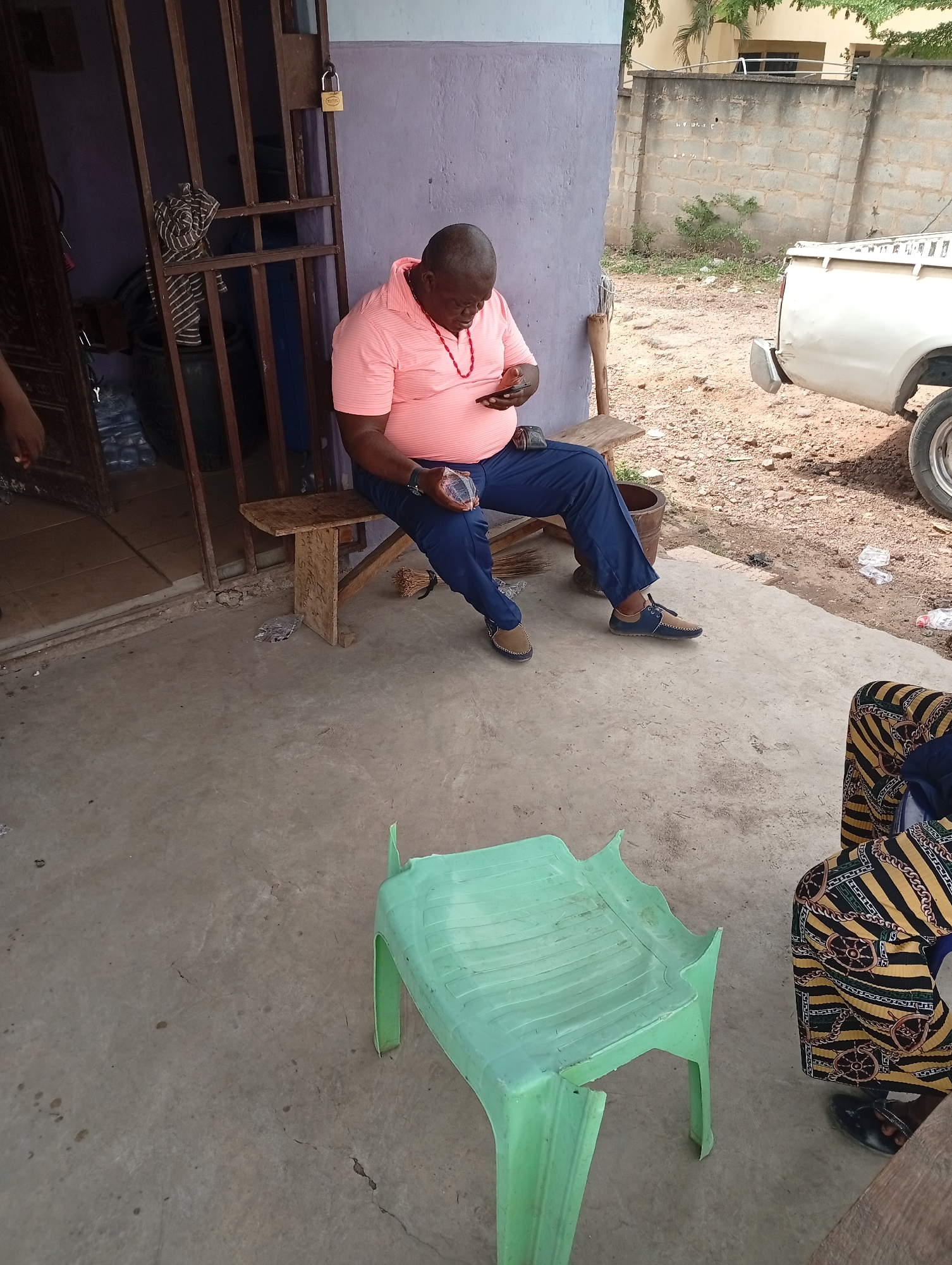 An outdoor scene depicts a man seated on a wooden bench, looking down at his hands or a device he is holding. He is wearing a pink short-sleeved polo shirt, blue trousers, and two-toned brown and blue shoes. A red beaded necklace is visible around his neck. To his left, part of another person is visible near a metal security gate, which has a brass padlock on it. The padlock shows "D&D" and "PADLOCK" text.
In the foreground, a light green plastic chair, possibly broken on one side, sits on a concrete floor. To the right, the leg of a third person is visible, wearing a black patterned fabric with gold and black chain-like designs.
The background features a dirt and gravel area, with a white pickup truck partially visible on the right. Beyond that, a grey concrete block wall and some green foliage and trees are present. The ground around the truck is littered with several plastic water bottles, one of which has "BIG Cola" visible on its label. A dark pot or container is seen near the metal gate. A bundle of what appears to be sticks, possibly a traditional broom, rests by the man's feet. The side of the wooden bench has some faint, carved or written text, but it is largely illegible. The overall setting suggests a casual, outdoor area, likely during the daytime under natural light.
