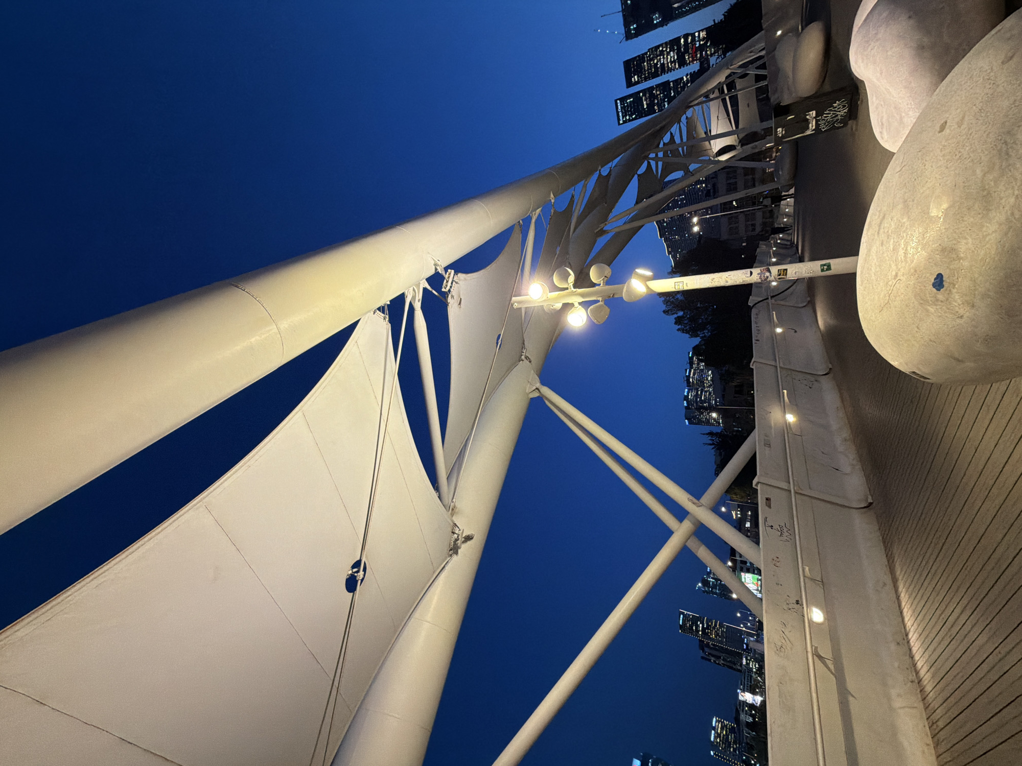 The image depicts the urban landscape of Tel Aviv, Israel, at night. The foreground features white, sail-like structures supported by metal poles, possibly part of a canopy or architectural installation. Adjacent to the structures is an elevated walkway with a wooden surface. In the background, a cluster of tall buildings punctuates the skyline, their lights illuminated against the deep blue, twilight sky. There are no people visible. Graffiti is observed on the side of the walkway structure. Light fixtures are attached to the metal structures, providing artificial illumination to the scene.