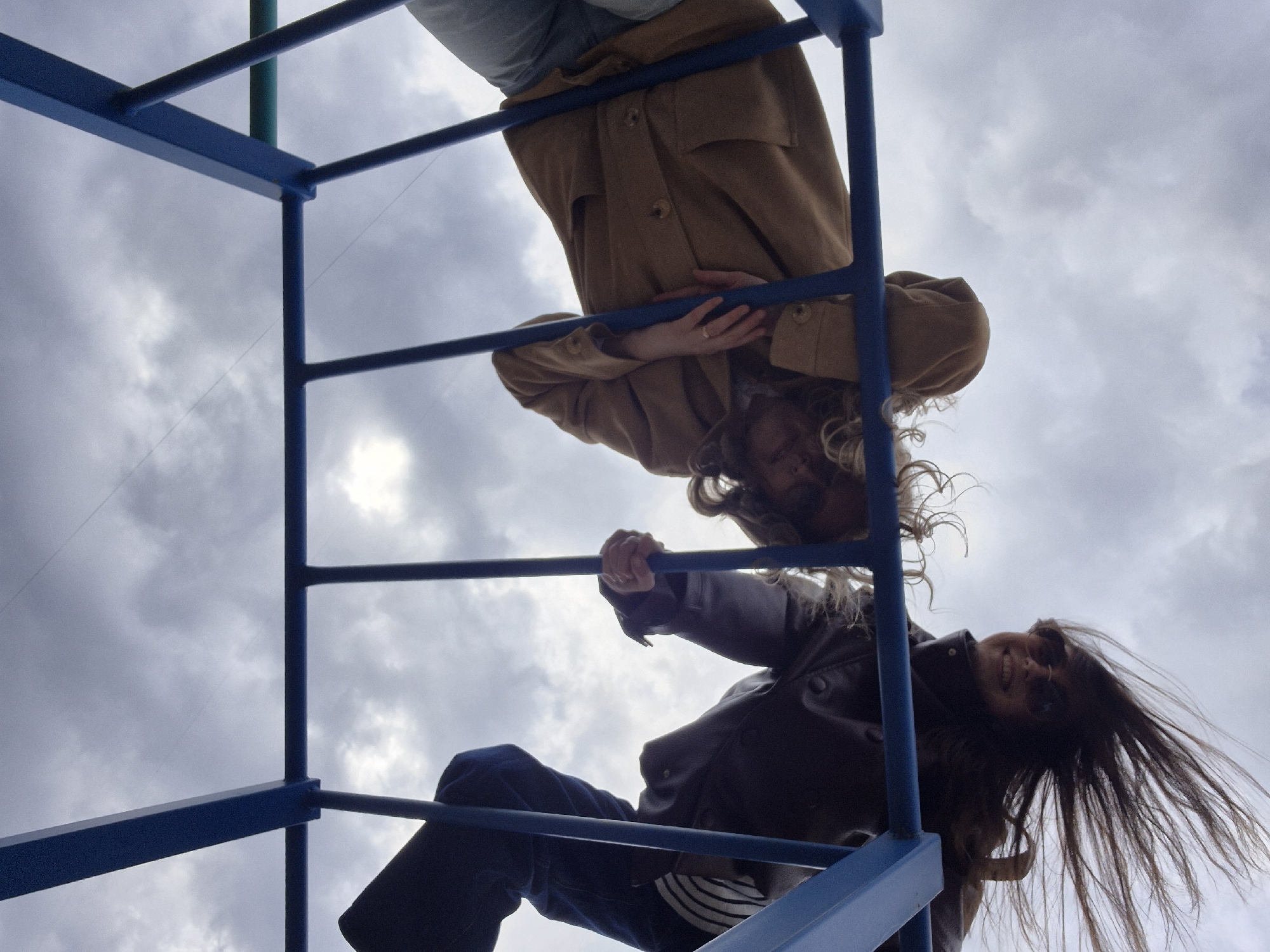 The image captures two individuals, likely young adults, hanging upside down from a blue metal climbing frame, viewed from a low-angle perspective looking upwards. The photo is oriented such that the subjects appear largely horizontal across the frame, with the cloudy sky forming the background.
The person positioned higher in the frame is wearing a light brown or tan-colored coat and light-colored trousers. Their hands are clasped together, and their light, curly hair hangs freely towards the bottom of the image.
Below them, the second person is clad in a dark jacket, dark pants, and appears to have a striped shirt visible beneath the jacket. This individual wears sunglasses and has dark, straight hair that is fanned out due to their inverted position. A clear smile is visible on their face, indicating enjoyment or playfulness as they grasp one of the climbing frame's bars.
The setting appears to be outdoors during daytime, under an overcast sky filled with soft, grey clouds. The blue metal structure of the climbing frame is prominent, forming a geometric pattern that frames the subjects. No specific location cues beyond the playground-like structure are discernible, and no text is visible in the image.