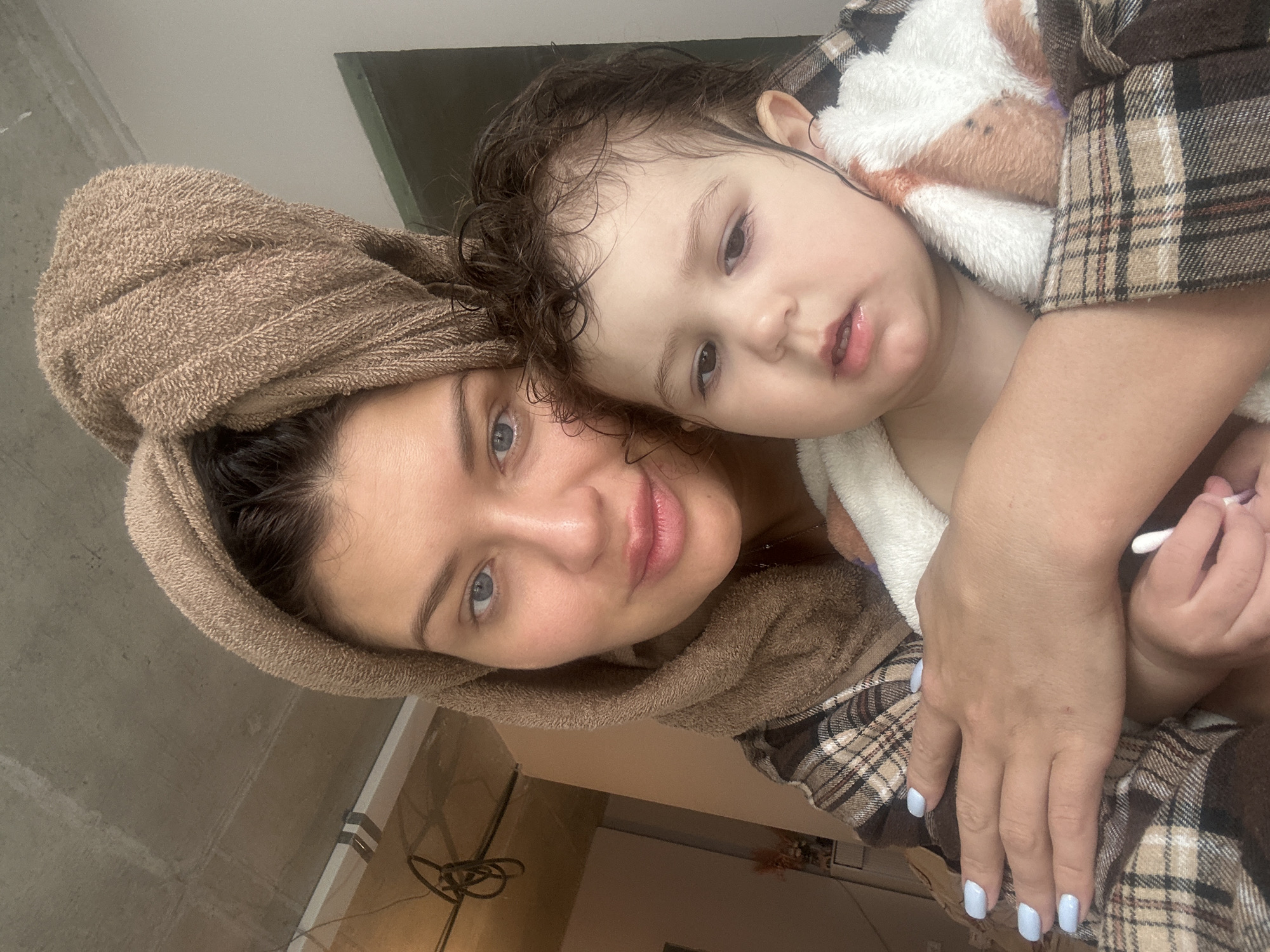 This close-up, rotated portrait features a woman and a young child in an intimate indoor setting, likely in Moscow, Russia. The woman, with bright blue eyes and a gentle expression, looks directly at the camera with a slight smile. Her dark hair is wrapped in a light brown towel, suggesting she has recently washed it or taken a bath. She is wearing a brown and white plaid shirt or robe, and her visible hand has light blue nail polish.
Leaning against the woman's head and shoulder is a young child, likely a toddler, with dark, damp, curly hair and curious dark eyes. The child also looks towards the camera. The child is partially covered by a soft, white and brown/orange plush robe, worn over or under a matching plaid garment. One of the child's hands is visible near the woman's hand, holding a white cotton swab.
The overall mood is tender and affectionate, capturing a moment of closeness between the two subjects, possibly after a bath or during a quiet, cozy time indoors. The background is simple and blurry, consisting of plain walls, offering no specific locational cues beyond the provided context. There is no visible text in the image.
