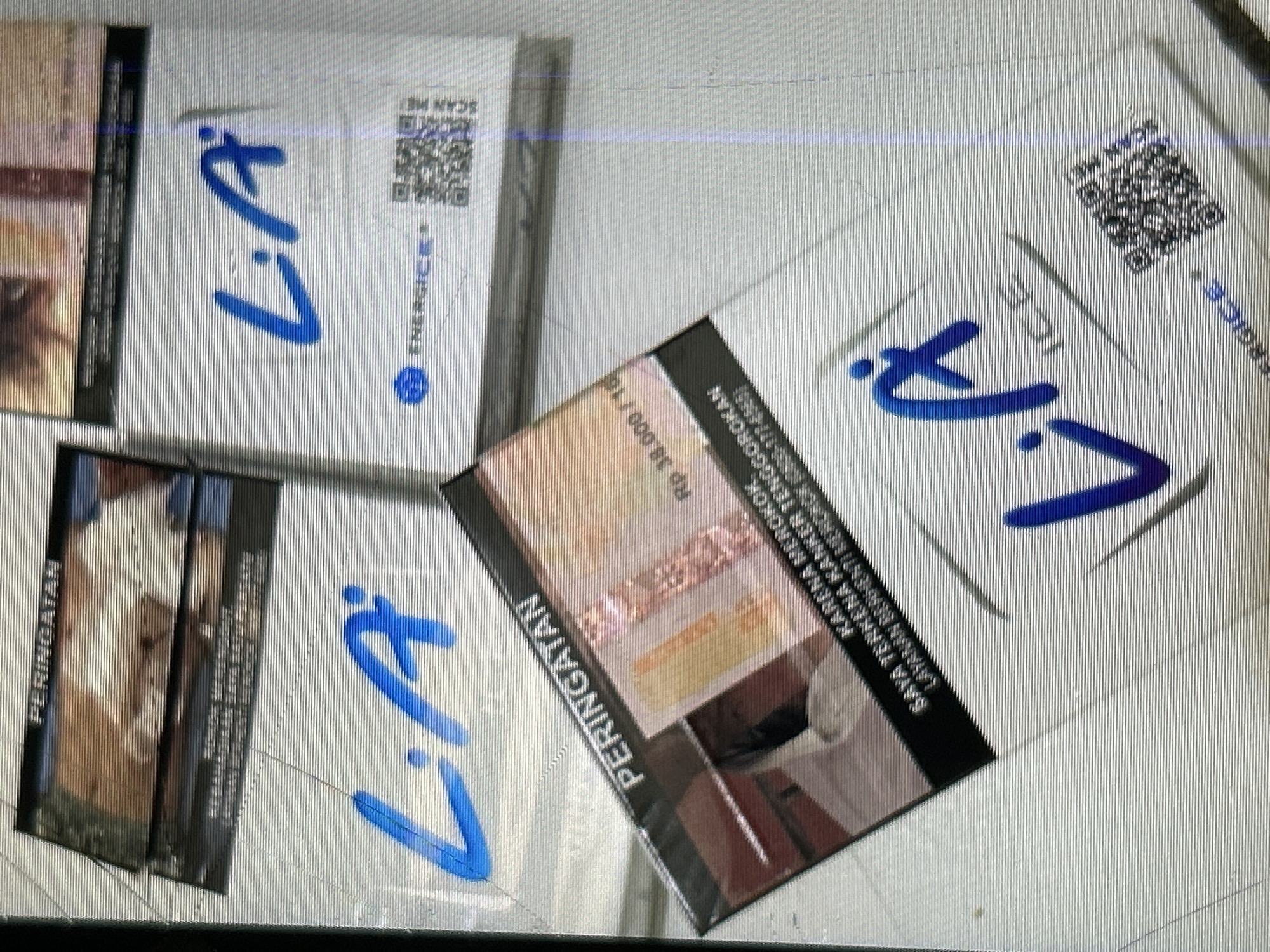 The image shows several packs of L.A. ICE cigarettes, some stacked and others slightly separated, on a textured surface that appears to be a table or counter. The background is out of focus but suggests an indoor setting. One pack in the foreground displays a warning label with text in Indonesian, "PERINGATAN" and "HARGA: Rp 38.000/10," indicating a price of Rp 38,000 for 10 cigarettes. Another pack in the lower right has the "L.A. ICE" logo and a QR code, along with the word "ENERGISE+". The lighting is somewhat dim, and the image quality is affected by horizontal lines, possibly due to the camera or scanning process. There are no people or animals visible in the image. The overall scene depicts a retail display or personal collection of cigarettes. The location in Depok, Indonesia, is not discernible from the visual elements present in the image.
