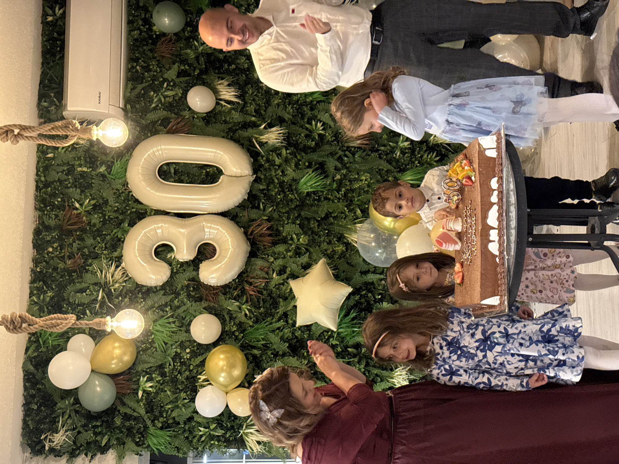 A group of people is gathered around a table in an indoor setting. The wall behind them is decorated with greenery, balloons, and inflatable number "30". There is an air conditioning unit on the upper left corner of the frame. A woman wearing a maroon dress is standing towards the bottom of the frame, taking a photo or video with her phone, looking towards the center. Four children are standing in front of a cake on the table. The cake appears to be chocolate. A man in a white shirt and dark pants is standing towards the top of the frame. The location is Buhovo, Bulgaria.