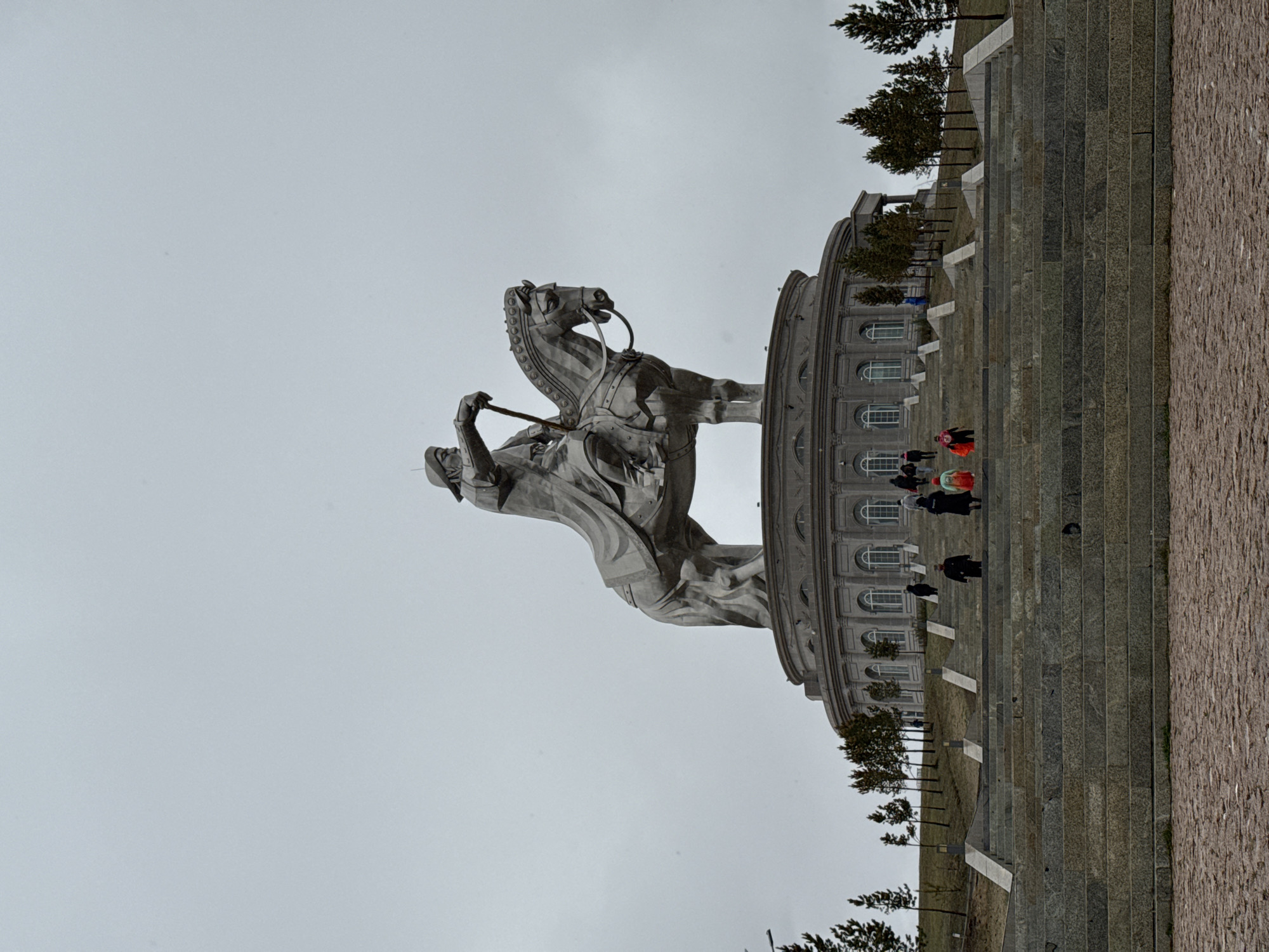 The image shows the Genghis Khan Equestrian Statue Complex located in Ulaanbaatar, Mongolia. The statue depicts Genghis Khan mounted on a horse. People are visible walking on a staircase leading up to the building behind the statue. The weather appears overcast, with a gray sky. Trees are visible around the staircase. The architectural style of the building is classical.
