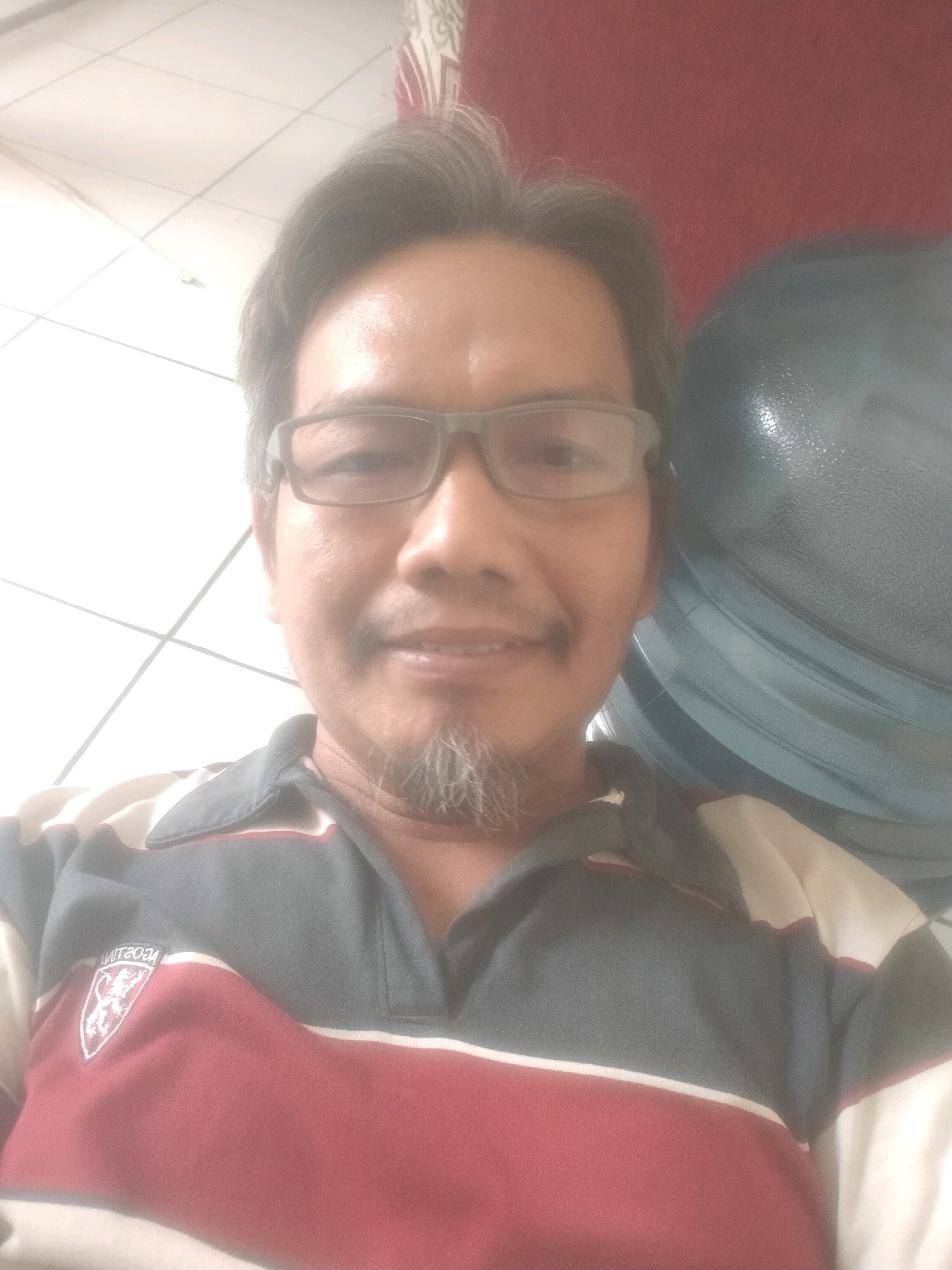 In Suka Sari, Indonesia, a man with glasses and a goatee is taking a selfie. He is wearing a striped polo shirt with a crest on the left side. Behind him, a large, blue water dispenser is visible, with a red wall in the background. The lighting suggests an indoor setting, possibly a home or office, with a tiled floor and ceiling. The overall impression is casual and relaxed.