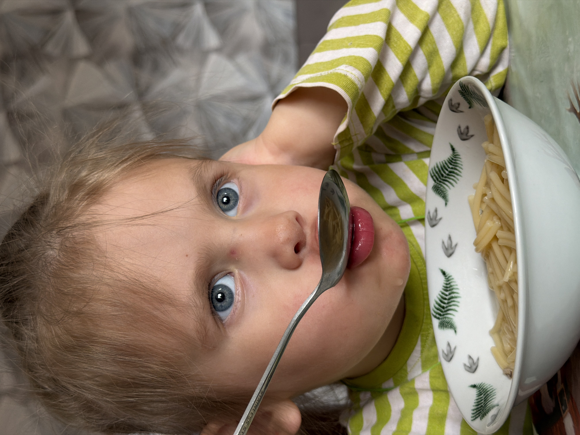 A young child with light blonde hair and striking blue eyes is depicted in a close-up, rotated portrait, looking directly at the viewer. The child appears to be a toddler, possibly between two and four years old. They are wearing a long-sleeved shirt with alternating green and white horizontal stripes.
The child is engaged in eating, or is about to eat, a meal. A silver spoon is positioned close to their mouth, with their tongue slightly visible. In the bottom right portion of the frame, a white bowl filled with plain, tubular pasta (resembling macaroni) is visible. The bowl features decorative elements, including green fern leaves and grey paw print motifs.
Notable details include the child's wide-eyed, focused gaze, conveying curiosity or concentration. A small amount of moisture or food residue is visible near the corner of the child's mouth. The spoon's handle clearly displays the engraved text "STAINLESS STEEL". The background is softly blurred, showing a grey, patterned texture, suggesting an indoor setting. The composition is taken from an angle that rotates the child so their head is on the left and their body extends towards the right, as if lying down or captured from above.
The image was taken in Chaikovsky, Russia.