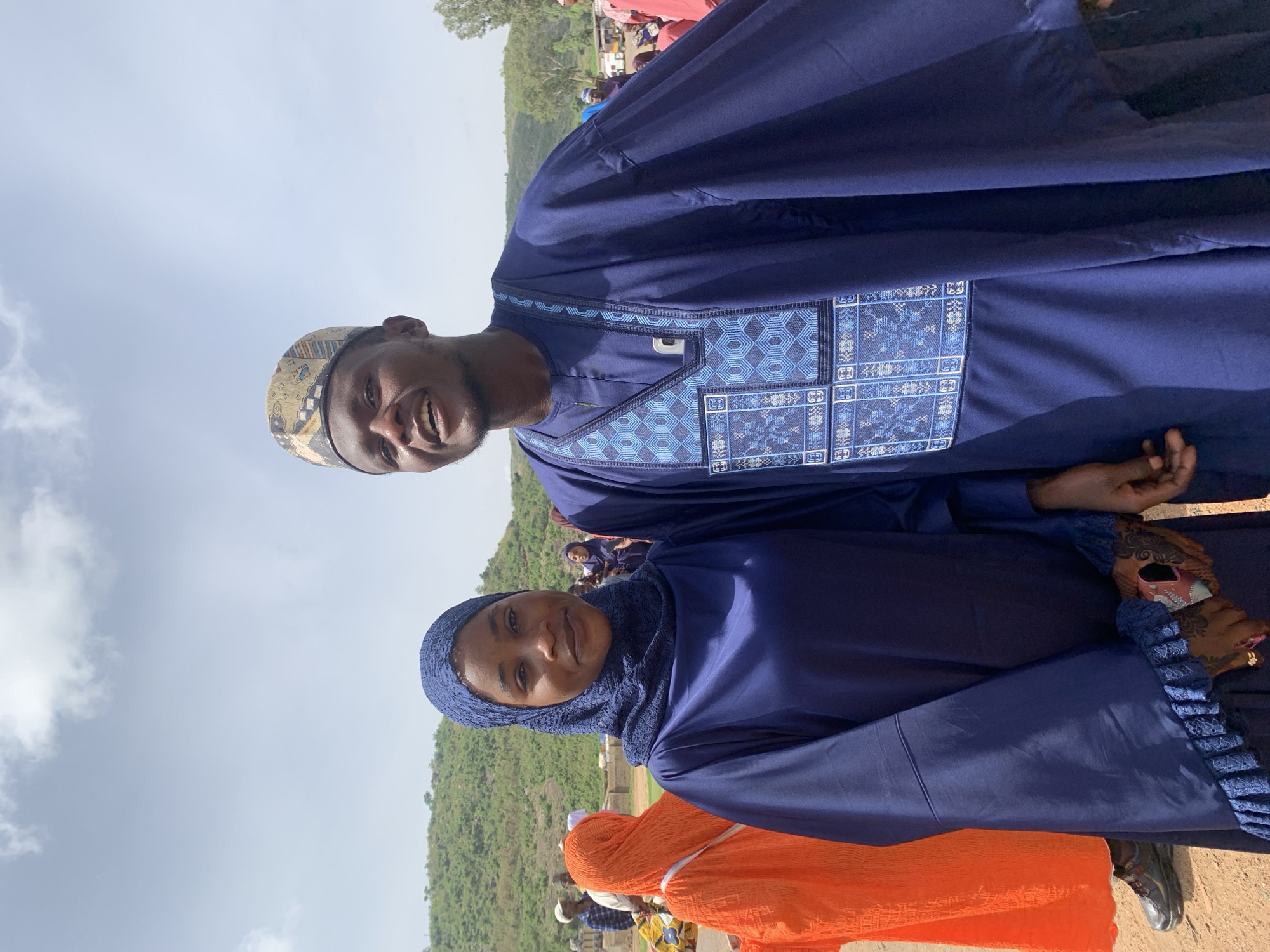 The image shows two people in what appears to be Naraguta Hausawa, Nigeria. One is a man wearing a blue traditional outfit with patterned embroidery, and a patterned hat. The other is a woman wearing a blue head covering and matching outfit. The setting appears to be outdoors, possibly at an event, with other people and some greenery in the background. The sky is overcast.