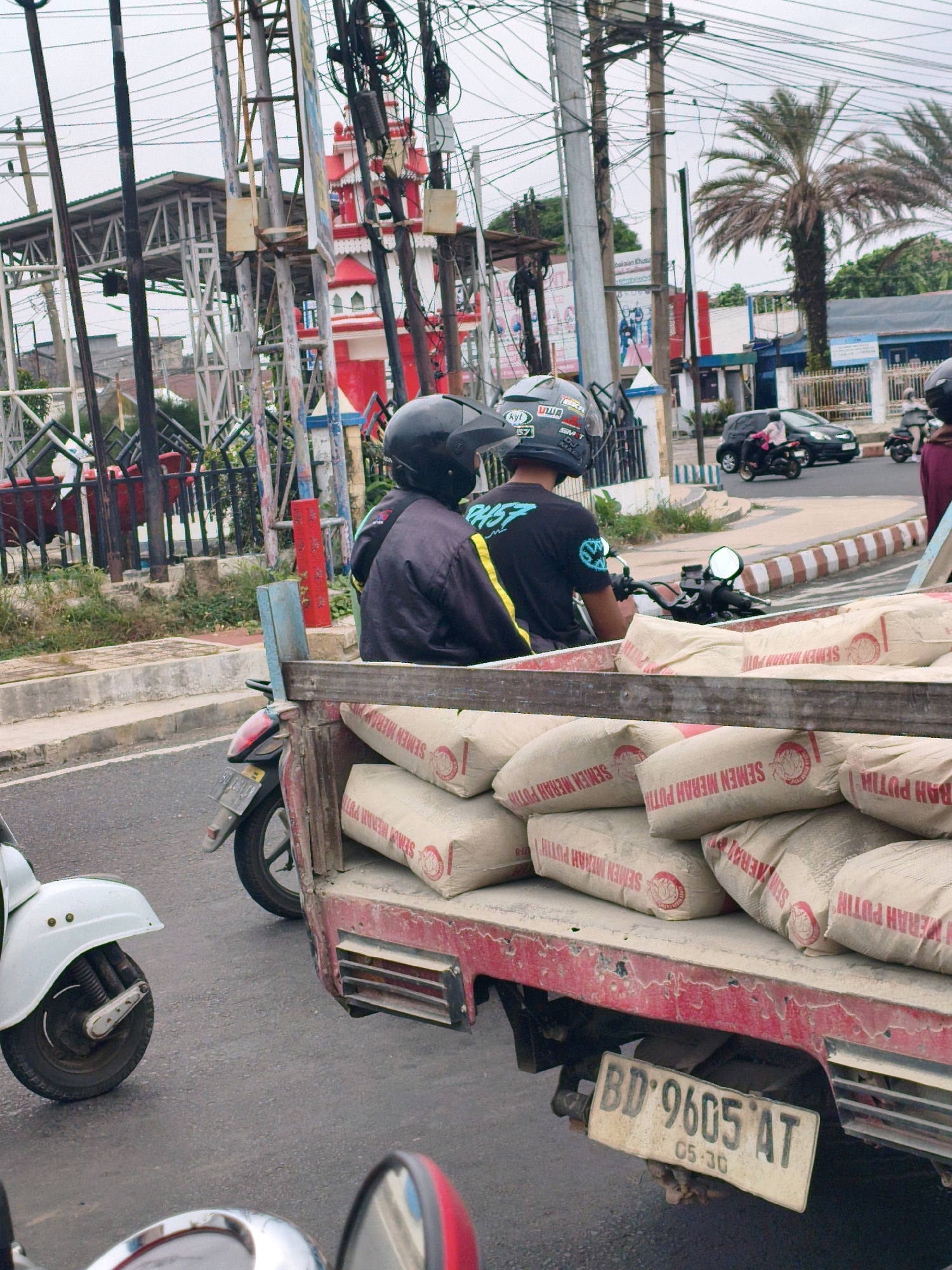 In Bengkulu, Indonesia, a motorbike with a trailer carrying several sacks of cement is parked on a street. The trailer is loaded with sacks labeled "SEMEN MERAH PUTIH" and "SEMEN MERAH PUTIH". A person wearing a black jacket and helmet is seated on the motorbike. Another person wearing a black jacket and helmet is also visible on a separate motorbike next to the trailer. In the background, a red and white building with traditional architecture is visible, along with utility poles and wires. Other vehicles, including a car and another motorbike, are on the street. The environment appears to be urban, with a cloudy sky. The license plate on the trailer reads "BD 9605 AT" with "05-30" below it.