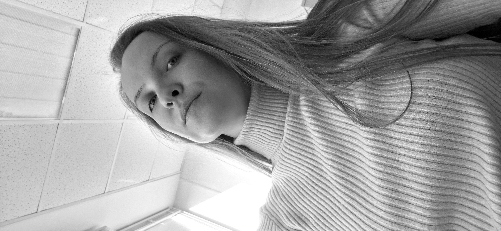 A low-angle, black and white photograph depicts a woman with long hair, wearing a ribbed turtleneck sweater. She is gazing down at the camera with a neutral expression. Behind her, there is a tiled wall and a suspended ceiling with fluorescent lights. The location is suggestive of a public restroom or similar institutional space. The location is not discernable to be Chaikovsky, Russia.