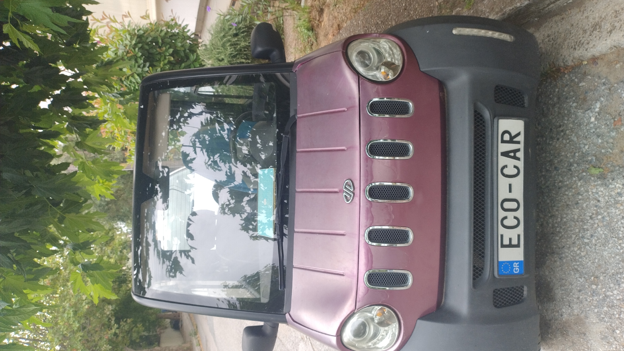 An outdoor, eye-level, front-facing shot captures a small, two-door, maroon colored electric car parked on a gravel surface. The vehicle has a black bumper with a European license plate displaying "GR ECO-CAR". The car's headlights are circular and prominent on either side of the front grille, which features five vertical, chrome-trimmed openings. Above the grille, a metallic logo is visible on the hood. The car's windshield reflects the surrounding environment, including trees with green leaves and a portion of the sky. The background shows foliage and hints of buildings, suggesting an urban or suburban setting. The car appears to be in good condition, parked at what is likely a roadside.