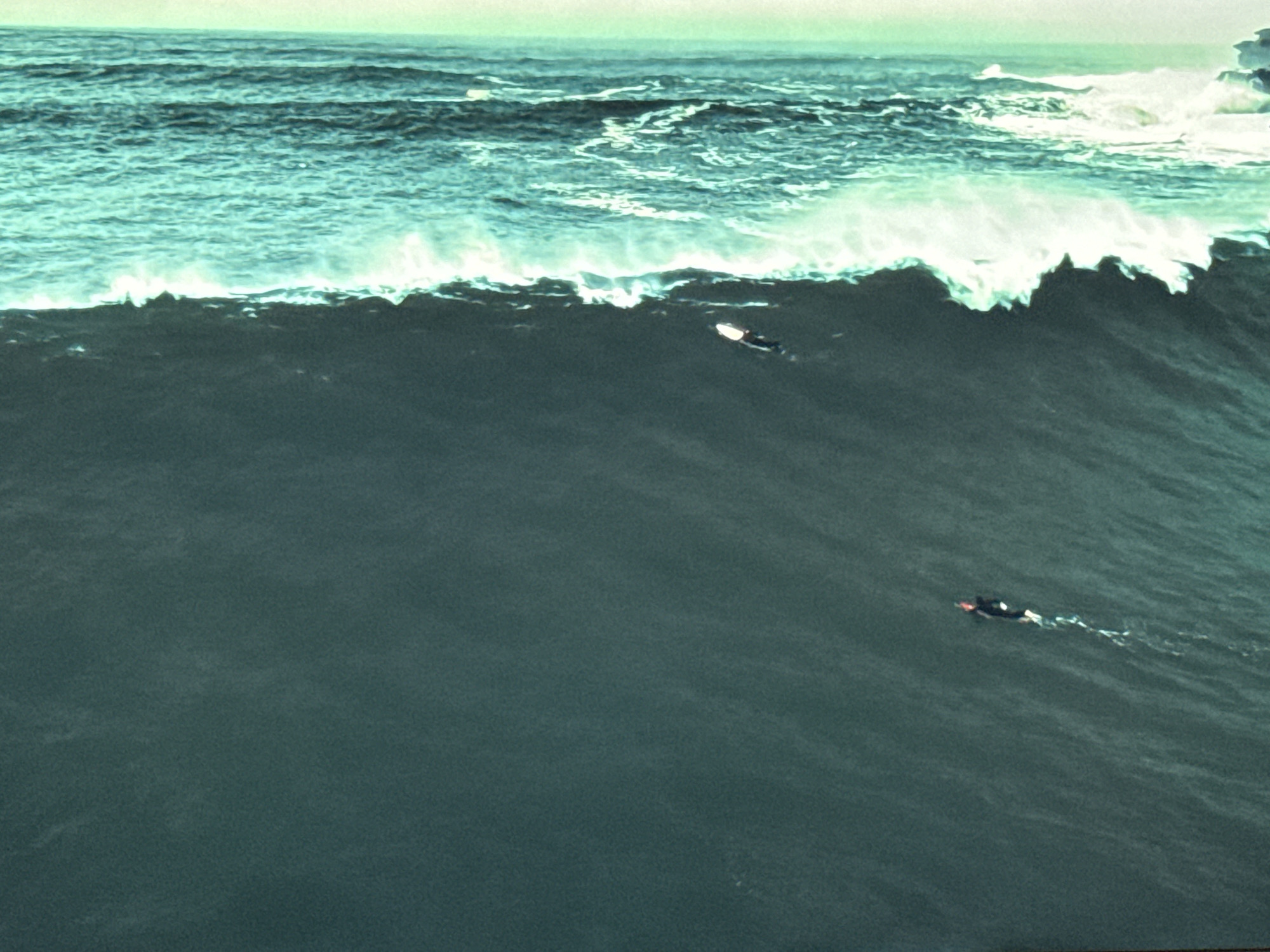An aerial, wide-angle shot captures two surfers in dark wetsuits navigating a massive, dark teal wave. The wave crests in the upper third of the image, a turbulent expanse of white foam against the deeper water. The first surfer is positioned on the right side of the image, about two-thirds of the way down the frame, appearing to be riding the wave. The second surfer is further left and higher up on the wave, nearer to the foamy crest. The scene is dominated by the immense scale of the ocean and the dark, powerful water. The sky above is a pale, muted green, hinting at overcast conditions. The location is identified as the city of Burgas, Bulgaria.