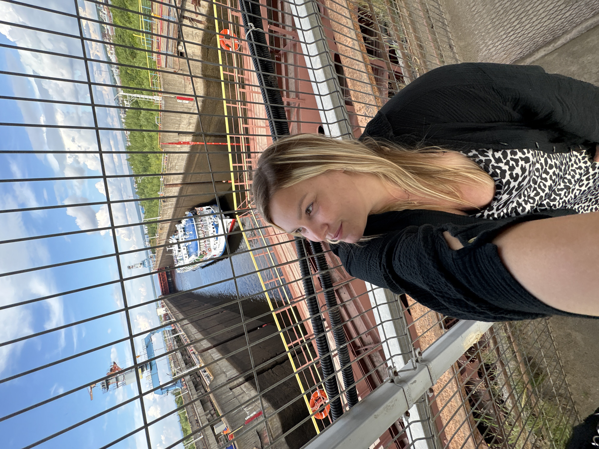 In this image, a woman with blonde hair is taking a selfie. She is wearing a black top and a leopard-print shirt underneath. She is leaning over a metal fence, with a large ship and a body of water visible behind her. The scene appears to be a lock or harbor, with industrial elements such as cranes and barges in the background. The sky is blue with some clouds, suggesting fair weather. The image is taken at an angle, with the woman in the foreground and the industrial landscape stretching out behind her. The overall impression is one of a casual moment captured while exploring a port or industrial area. There is no visible text in the image.