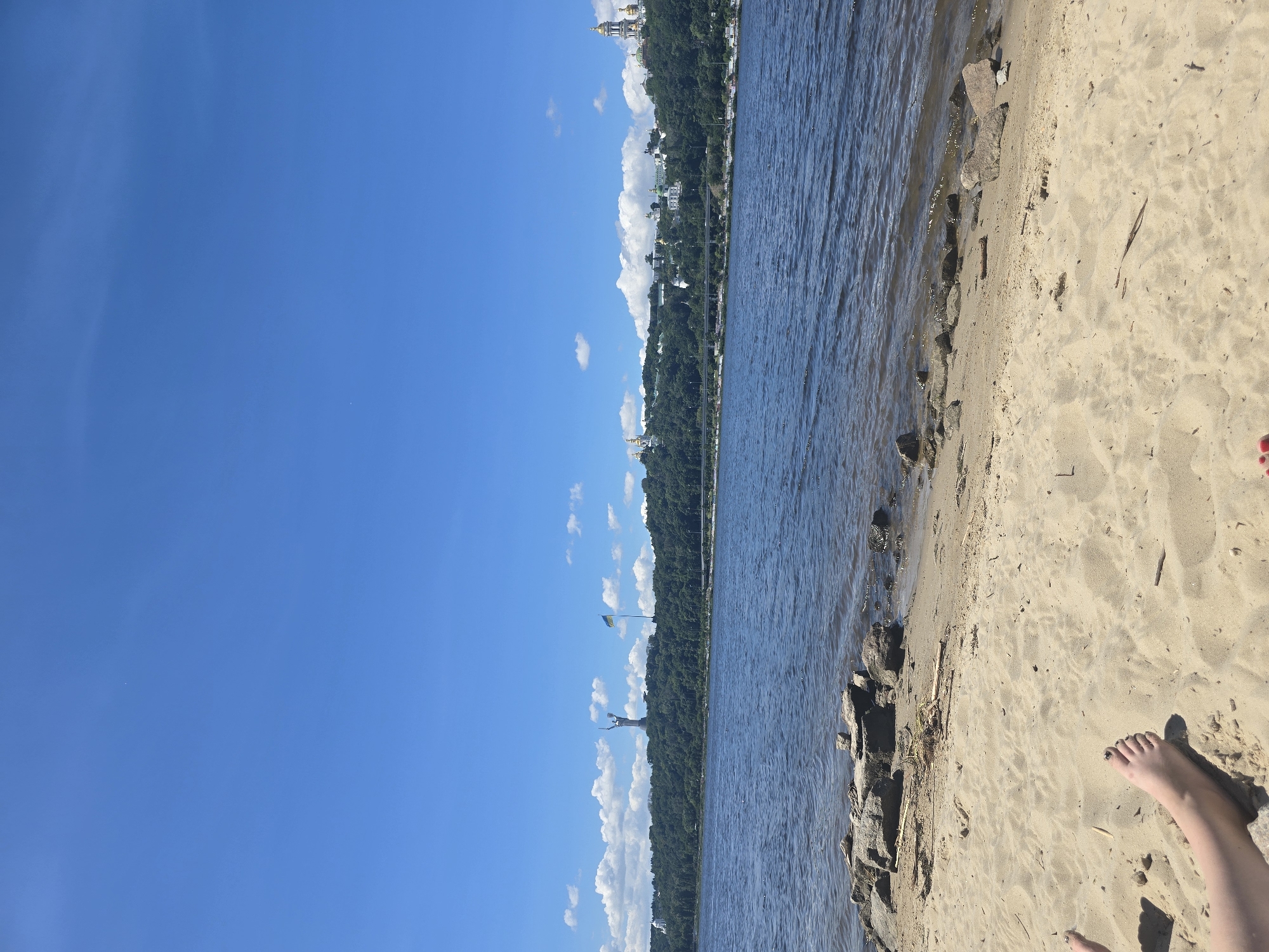 The image captures a serene view of the Dnieper River in Kyiv, Ukraine, from a sandy beach. In the foreground, a person's bare feet are visible on the sand, suggesting a leisurely moment. The water of the river is calm, reflecting the bright blue sky dotted with scattered white clouds.
Across the river, the iconic silhouette of the Motherland Monument stands prominently on the hill, a powerful symbol of the city. Beside it, the Ukrainian flag flutters in the breeze. Further along the skyline, the golden domes of Kyiv Pechersk Lavra, a historic monastery complex, gleam under the sun, adding to the city's spiritual and architectural grandeur. The lush green trees covering the hills provide a verdant backdrop to these landmarks. A bridge is visible in the middle ground, spanning the river. The scene depicts a peaceful day by the water, offering a glimpse of Kyiv's prominent landmarks.