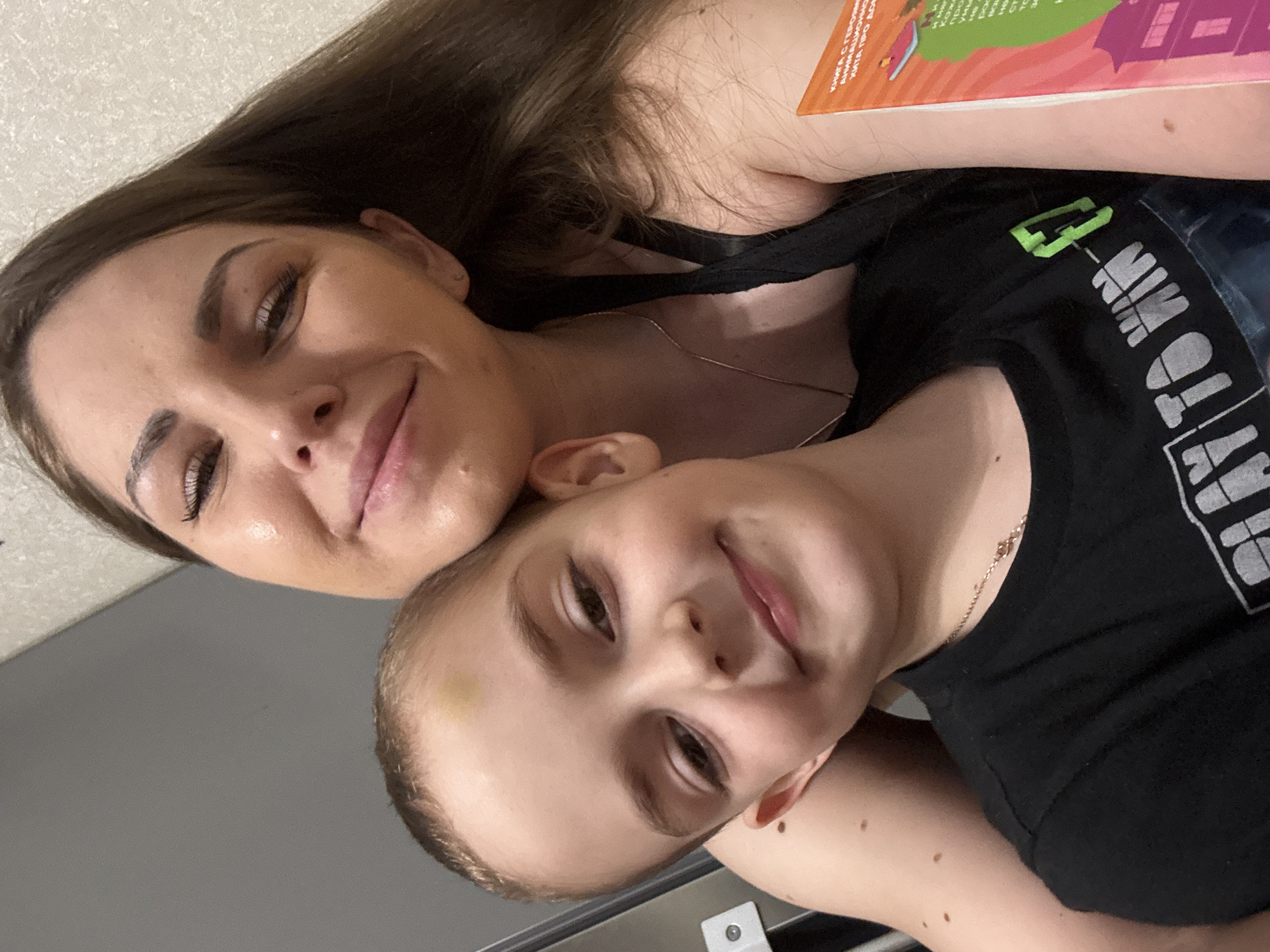 A selfie image shows an adult and a child posing closely together. The adult, likely a female, has long brown hair, fair skin, and wears a black tank top with the partially visible phrase "PLAY TO WIN." A book with colorful illustrations is partially visible above her head. The child has light brown hair, fair skin, and also wears a black shirt. A small bruise is visible on the child's forehead. Both are smiling slightly. The background appears to be a simple wall and a gray surface. The specific location, Раздолье, Russia, cannot be definitively determined from the image alone.