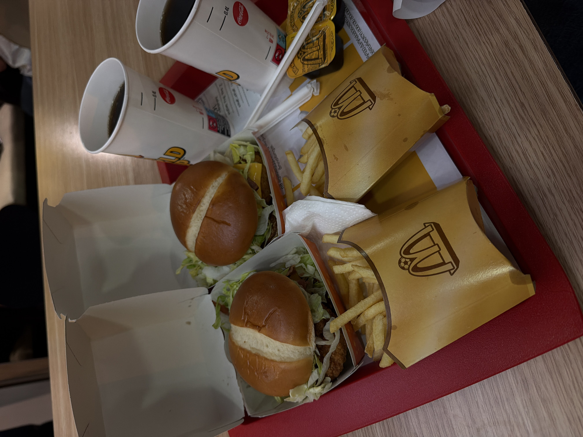 The image shows two McDonald's meals on a red plastic tray placed on a wooden table. Each meal consists of a burger in a white paper box, a carton of french fries, and a Coca-Cola cup. The burgers are on buns with lettuce. The french fry cartons are gold colored with a McDonald's logo and a soccer ball design. One of the Coca-Cola cups has a label that includes "0.4L" and a Coca-Cola logo. There are also two small packets of sauce, likely ketchup or mayonnaise, with McDonald's branding on them, and two white straws. The background is blurred but appears to be an indoor setting, possibly a McDonald's restaurant. The image quality is slightly grainy, with a warm, artificial light. There are no people clearly visible in the image, only parts of what appear to be individuals in the blurred background. The scene suggests a casual meal, possibly a takeaway order. The presence of McDonald's branding and the specific cups and packaging are indicative of this fast-food chain. The mention of Tbilisi, Georgia, in the prompt is not supported by any visual evidence within the image itself.