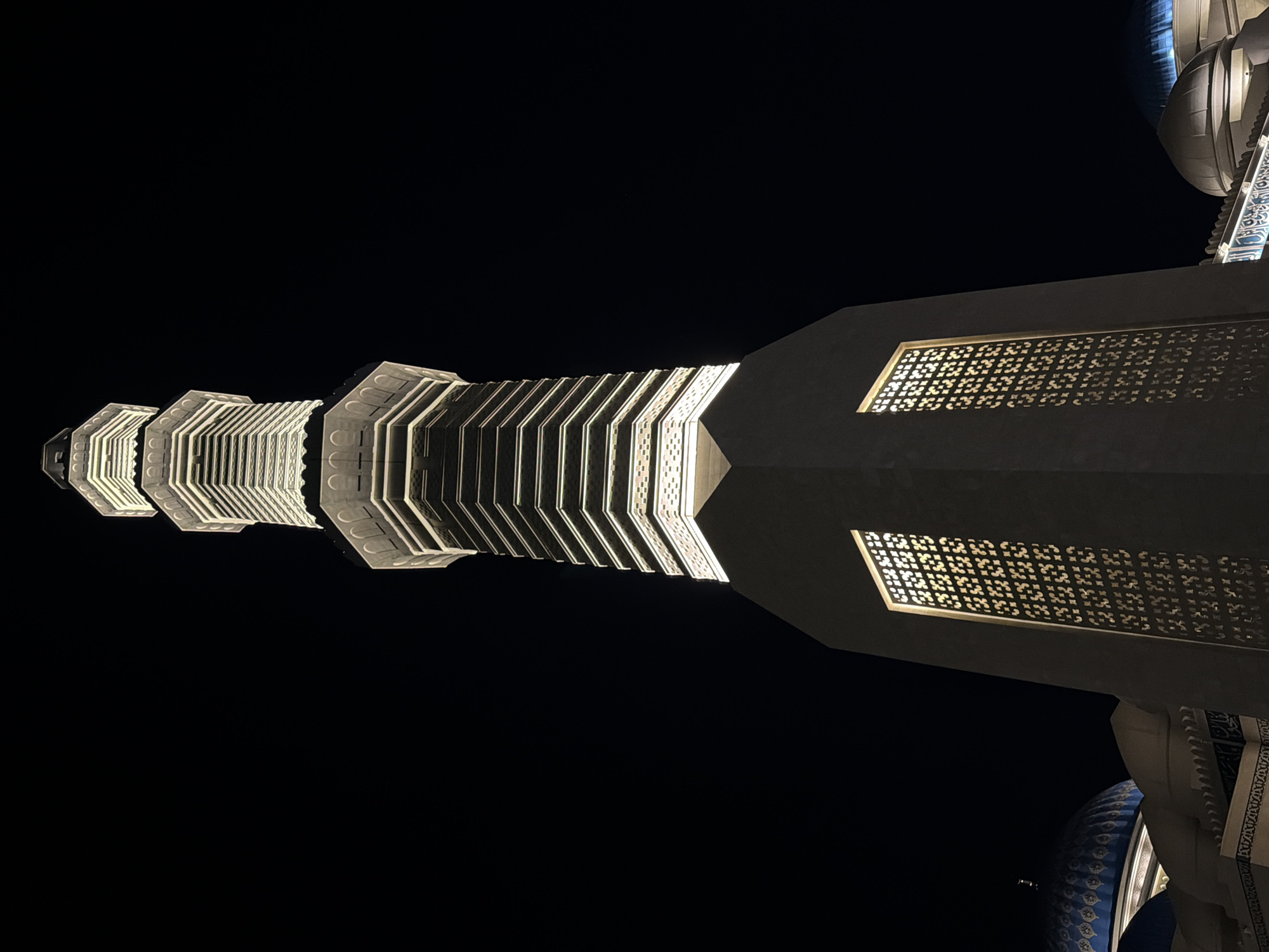 This is a low-angle, night-time shot of a minaret, a tall, slender tower, that is part of the Hazrat Sultan Mosque in Astana, Kazakhstan. The minaret is illuminated by warm, white lights that highlight its layered, octagonal structure and intricate architectural details. Two large windows with ornamental grills are visible on the base of the minaret, casting illuminated patterns on the surrounding darkness. To the left and right of the minaret, parts of the mosque's blue domes and other architectural elements are visible. The sky is completely dark, indicating it is nighttime. There are no people visible in the image.