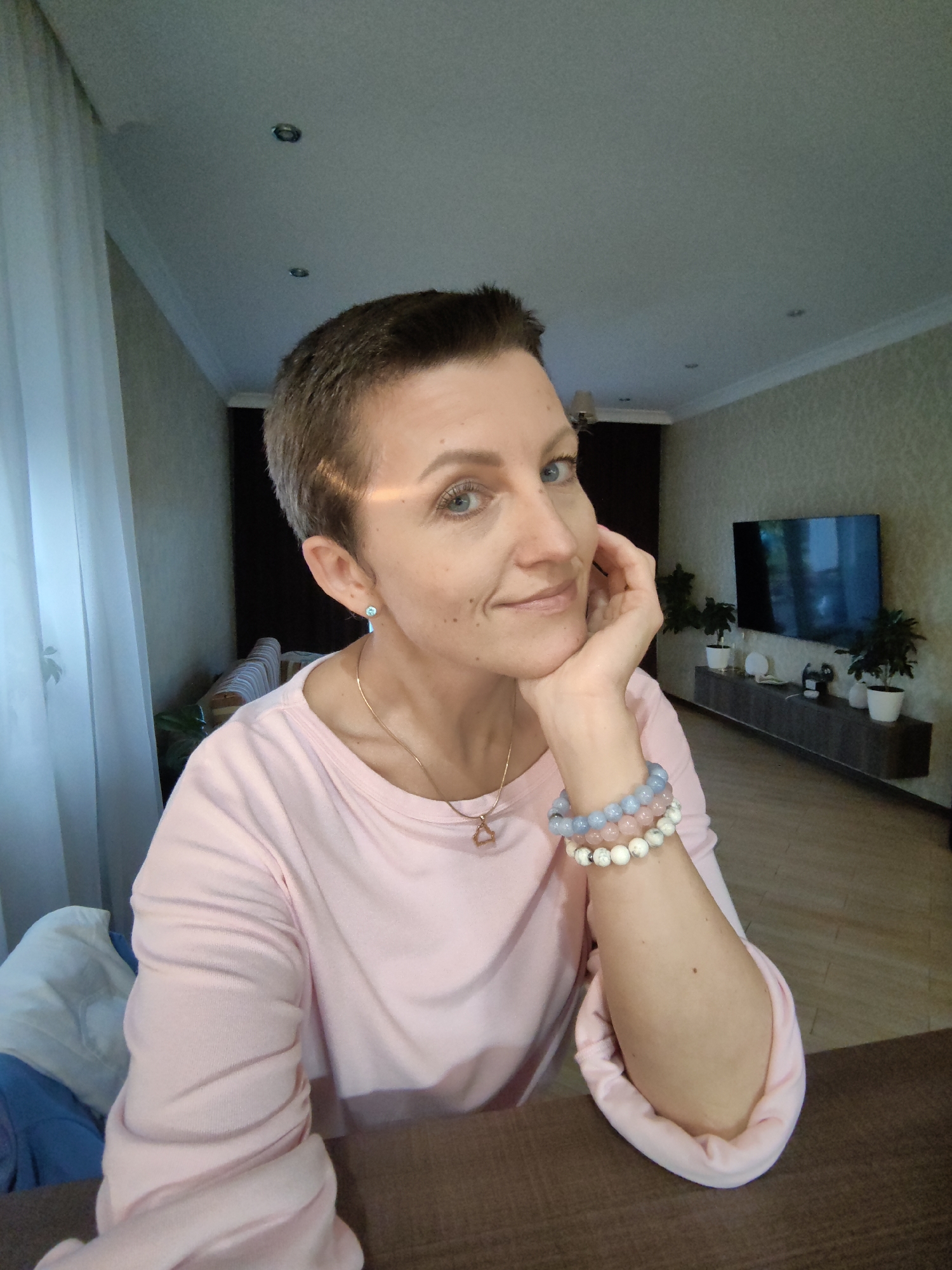 A close-up, selfie-style shot shows a woman with short brown hair and blue eyes in an indoor setting. She is wearing a pale pink long-sleeved shirt and resting her chin on her hand. Three beaded bracelets are visible on her left wrist. In the background, a television is mounted on a wall unit, with several potted plants placed on and around it. A sheer white curtain is visible to the left of the woman, and a couch with striped cushions can be seen in the distance. The lighting suggests an indoor environment. This image was taken in Stowbtsy, Belarus.