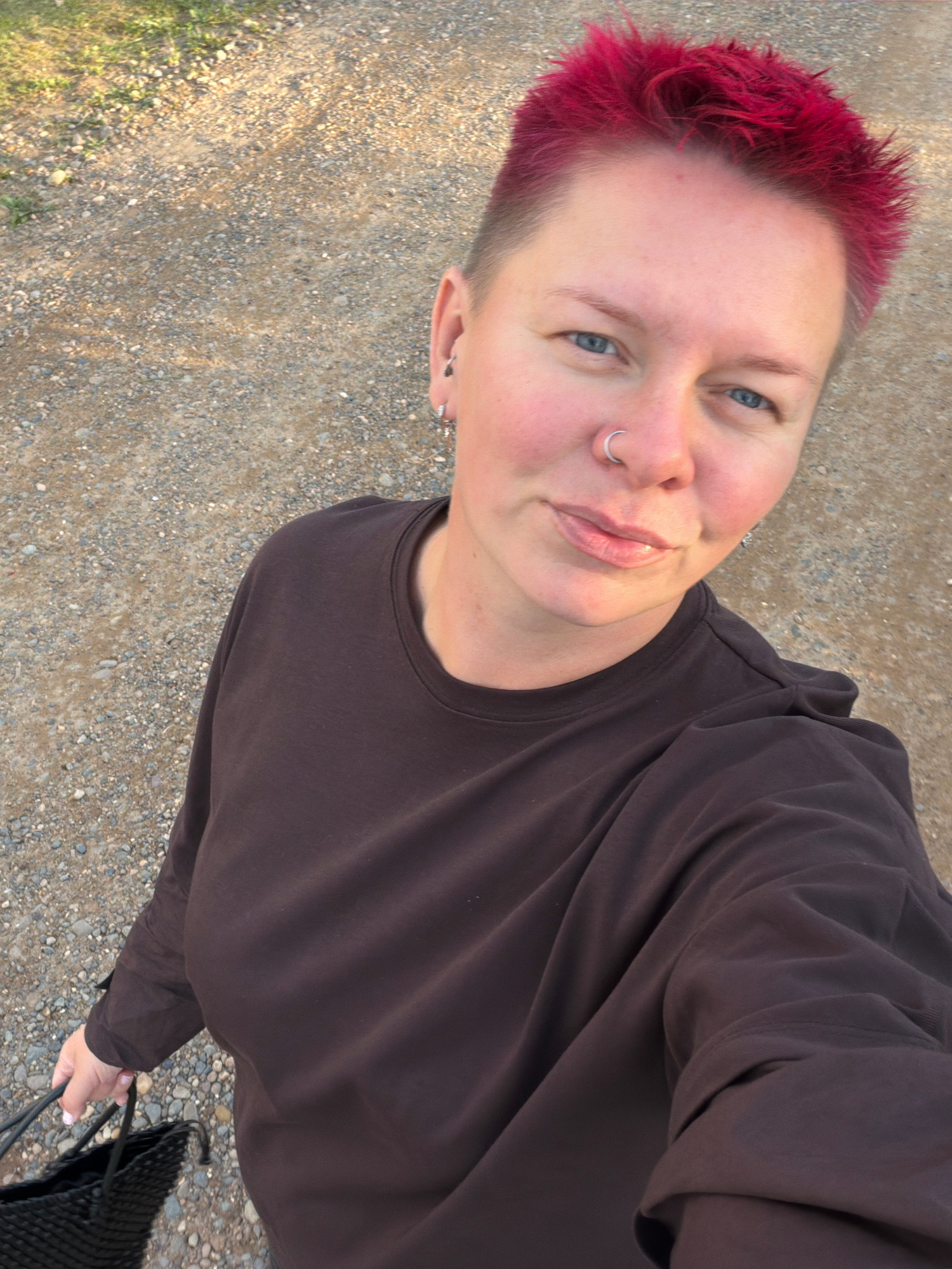 The image shows a person with short, bright pink hair and a nose ring, wearing a dark brown long-sleeved shirt. They are holding a black woven bag in their left hand. The ground beneath them appears to be gravel and dirt, with some patches of green vegetation in the background. The lighting suggests it is daytime, possibly late afternoon or early morning due to the long shadows. There is no identifiable information in the image to indicate a specific location within the city of Chaikovsky, Russia.