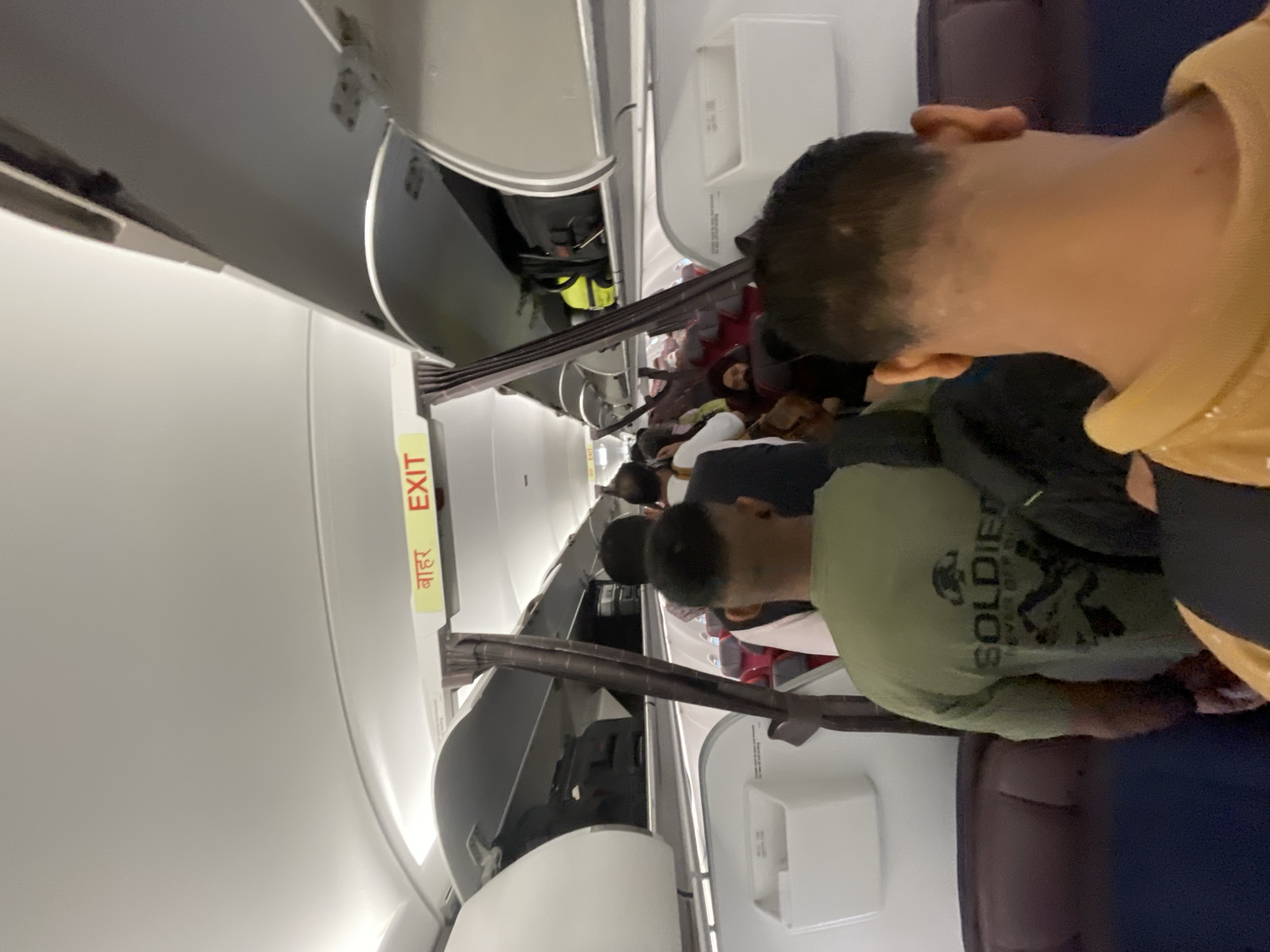 The image shows the interior of an airplane cabin, with passengers standing in the aisle, seemingly queuing. The overhead compartments are open, revealing luggage. An "EXIT" sign, in both Hindi and English, is visible above the aisle. The seats appear to be upholstered in a dark color, possibly red or maroon, and there are white plastic bins attached to the walls. The lighting is artificial and diffused, typical of an airplane cabin. The location of Raipur Khurd, India, is not directly indicated by any visual cues in the image itself. The people in the image are facing away from the camera, with their backs visible as they move down the aisle. One person in the foreground wears a green t-shirt with the word "SOLDIER" printed on it.