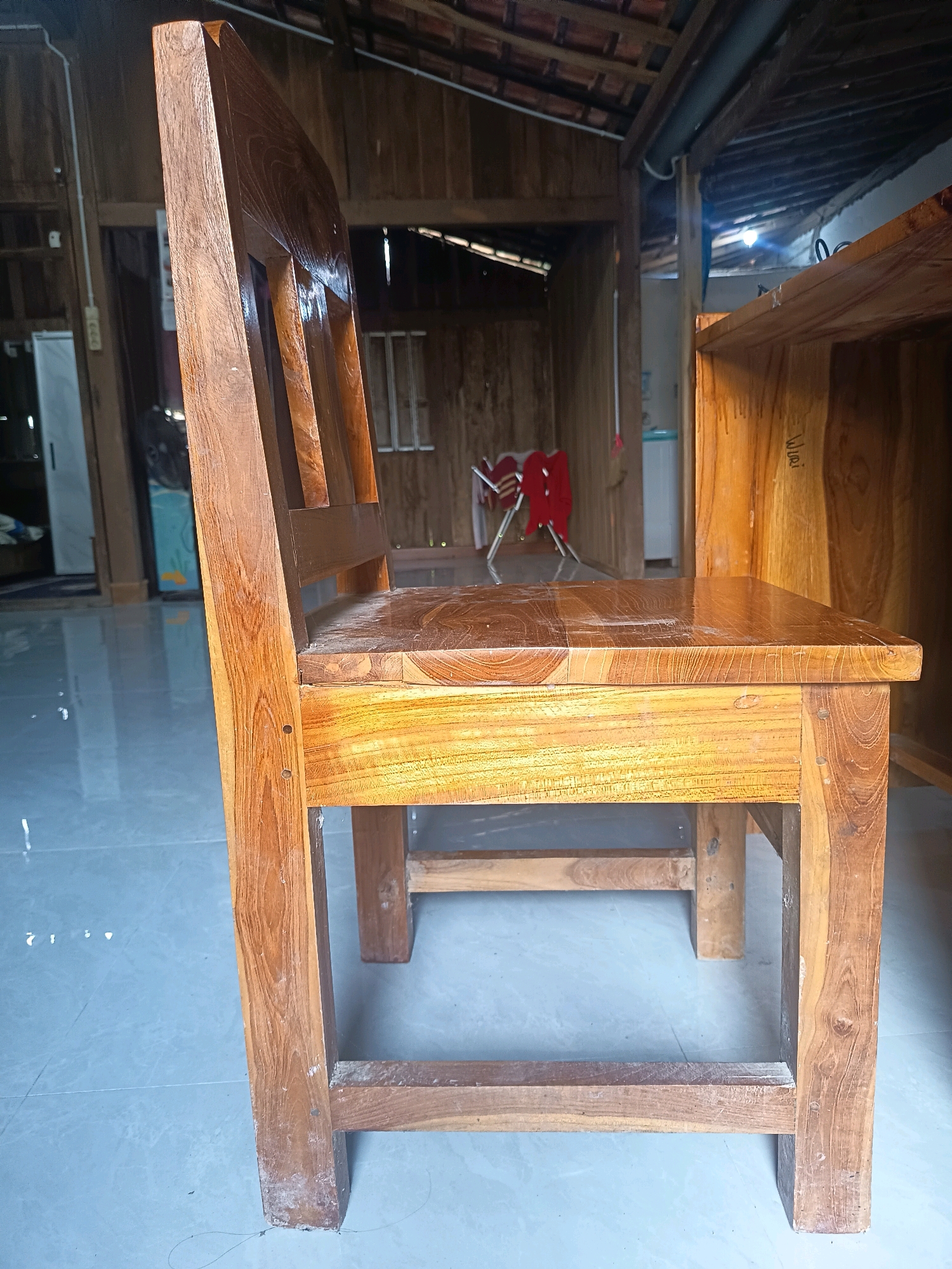 The image depicts a side profile of a wooden chair. The chair is made of light brown wood with a visible grain pattern. It has a simple design with a solid seat and a backrest composed of several vertical slats. The chair appears to be indoors, on a light-colored tiled floor. In the background, a wooden structure is visible, suggesting it might be a home or workshop. The lighting is adequate, casting shadows that highlight the texture of the wood. The overall scene is unremarkable, and there is no indication of any specific event or activity. The location of Sidorejo, Indonesia, is mentioned, but there are no visual cues in the image to confirm or deny this specific location. The chair is the primary subject, and its construction and material are the most notable features.
